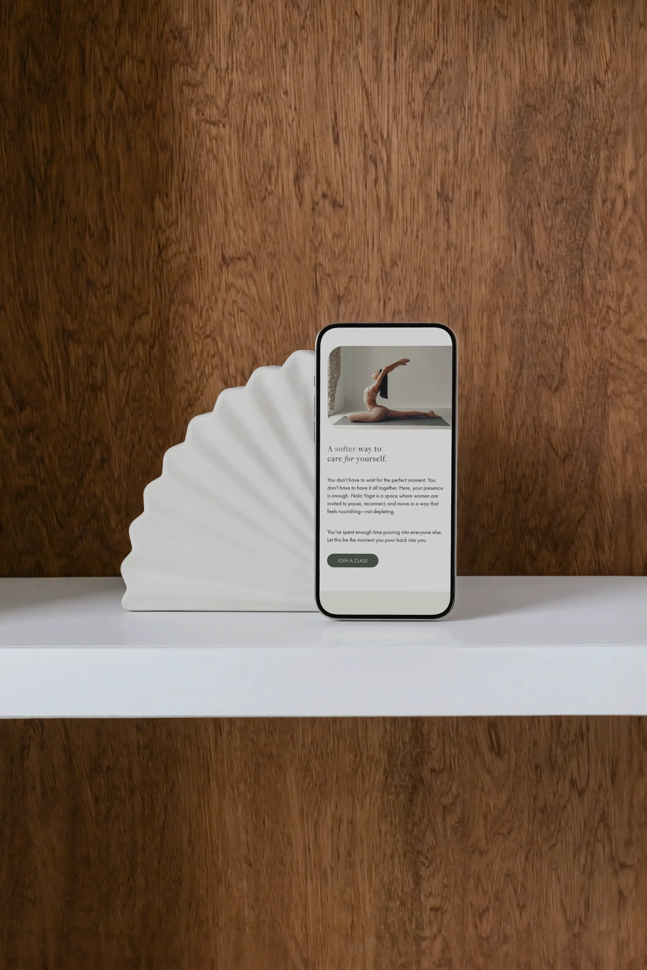 A smartphone displaying a website with an image of a woman performing a yoga pose on a mat, next to a white decorative object with a wavy edge, on a white surface with a wooden background.