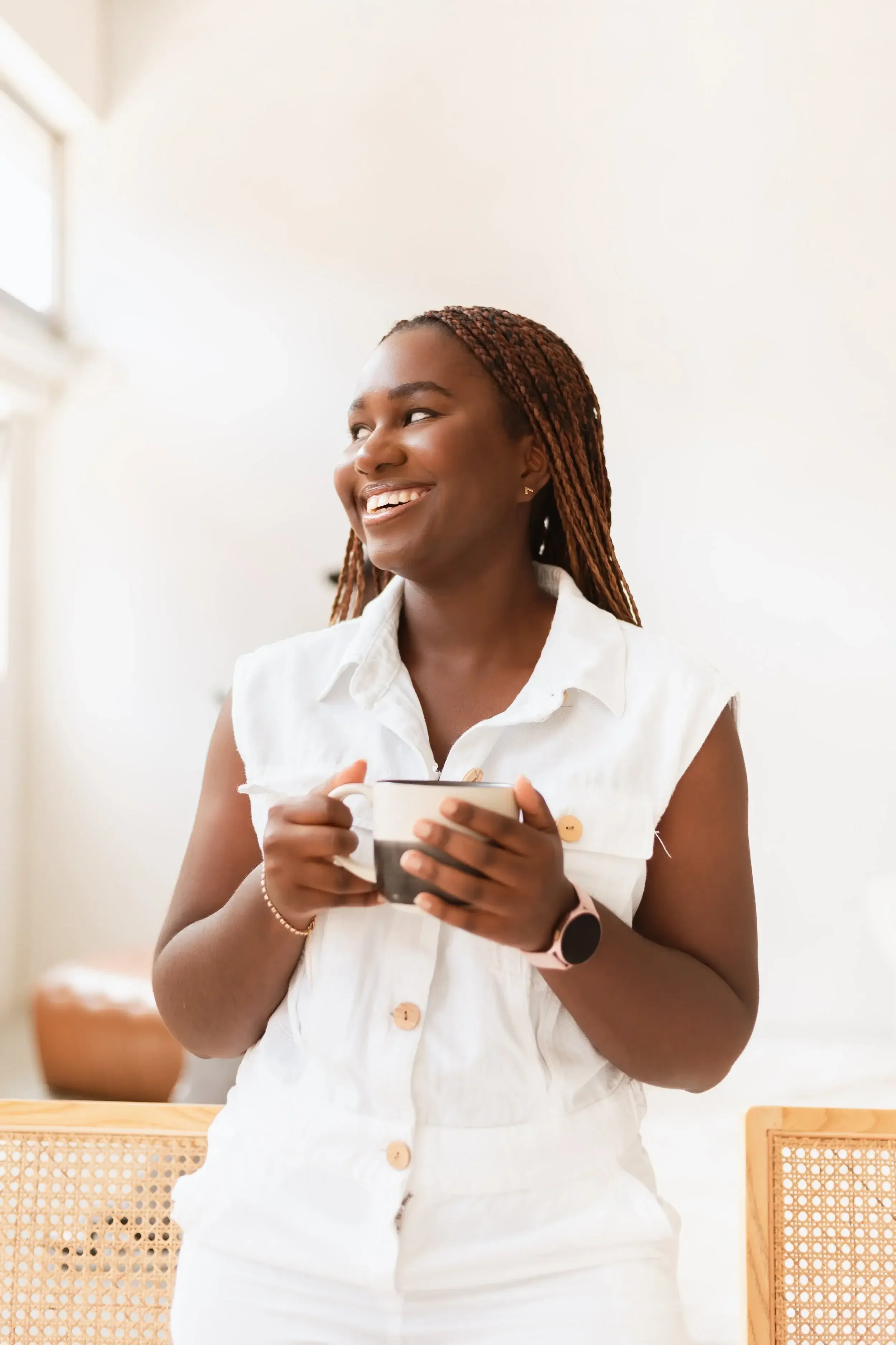 A woman with braided hair wearing a sleeveless white button-up dress, smiling and holding a mug, sitting on a wooden chair in a bright room.