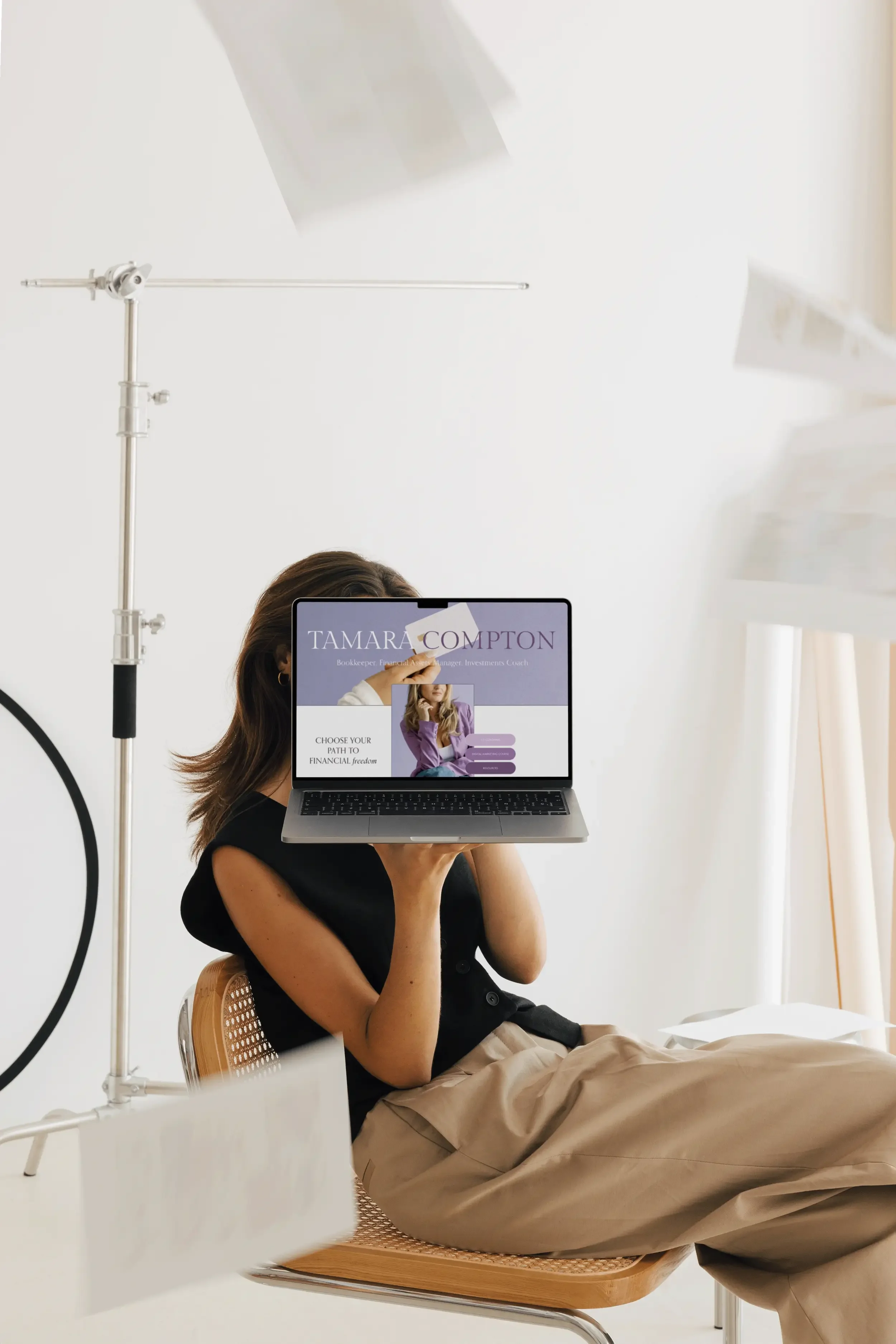 A woman sitting on a chair holding a laptop in front of her with the screen displaying a website for Tamara Compton, a financial advisor. She is in a bright room with white walls, surrounded by photography equipment and papers.