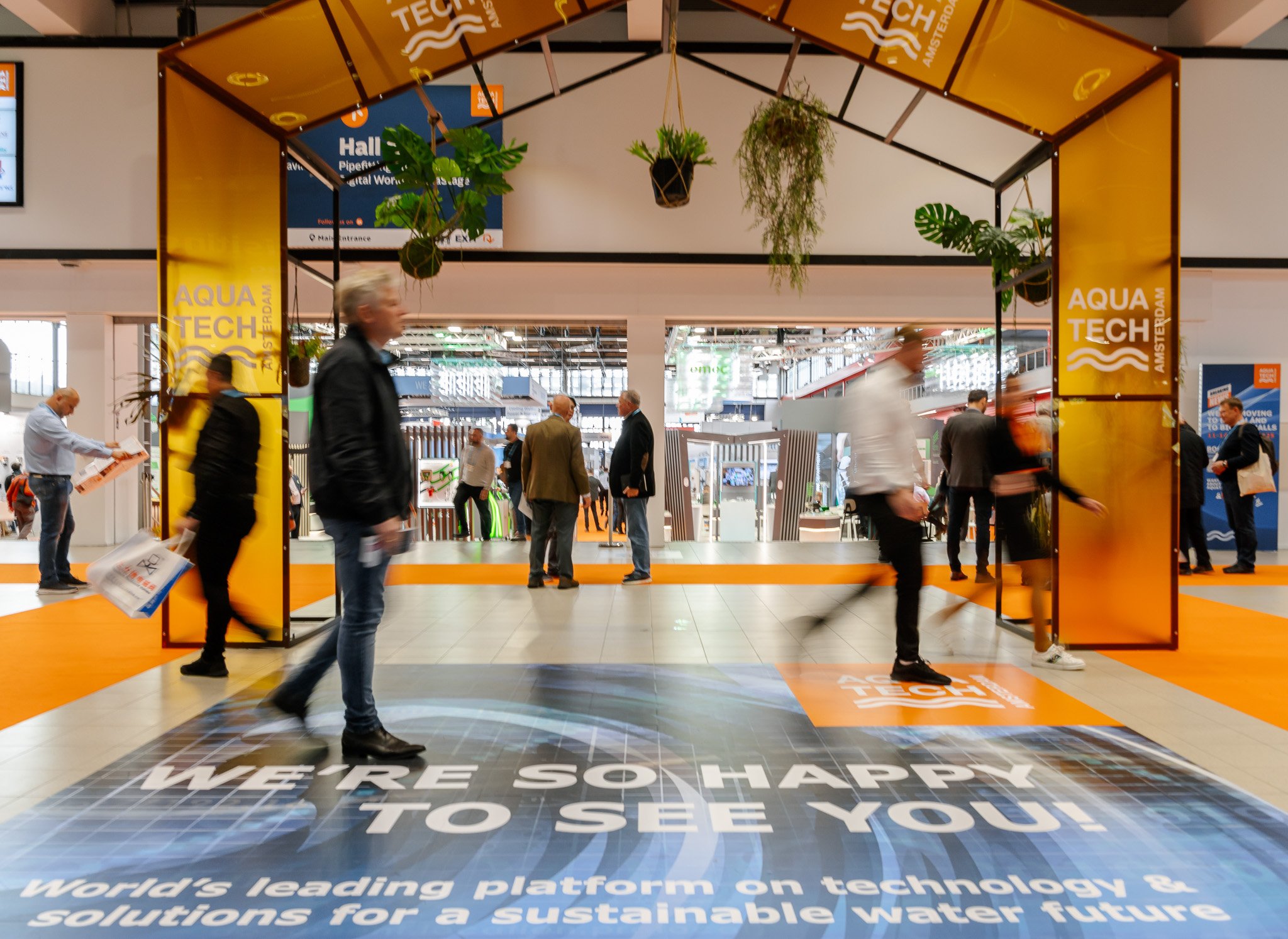 Eingang zur Aqua Tech Messe mit mehreren Leuten, die sich im Bereich bewegen. Im Vordergrund ein Bodenaufkleber mit der Aufschrift 'WE'RE SO HAPPY TO SEE YOU!' und weiteren Text über technologische Wasserlösungen. Das Messegelände ist modern mit grünen Pflanzen und einer großen Ausstellungshalle im Hintergrund.