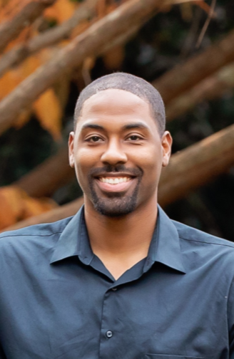 A smiling man with a short beard and mustache wearing a navy blue button-up shirt, standing outdoors with tree branches in the background.