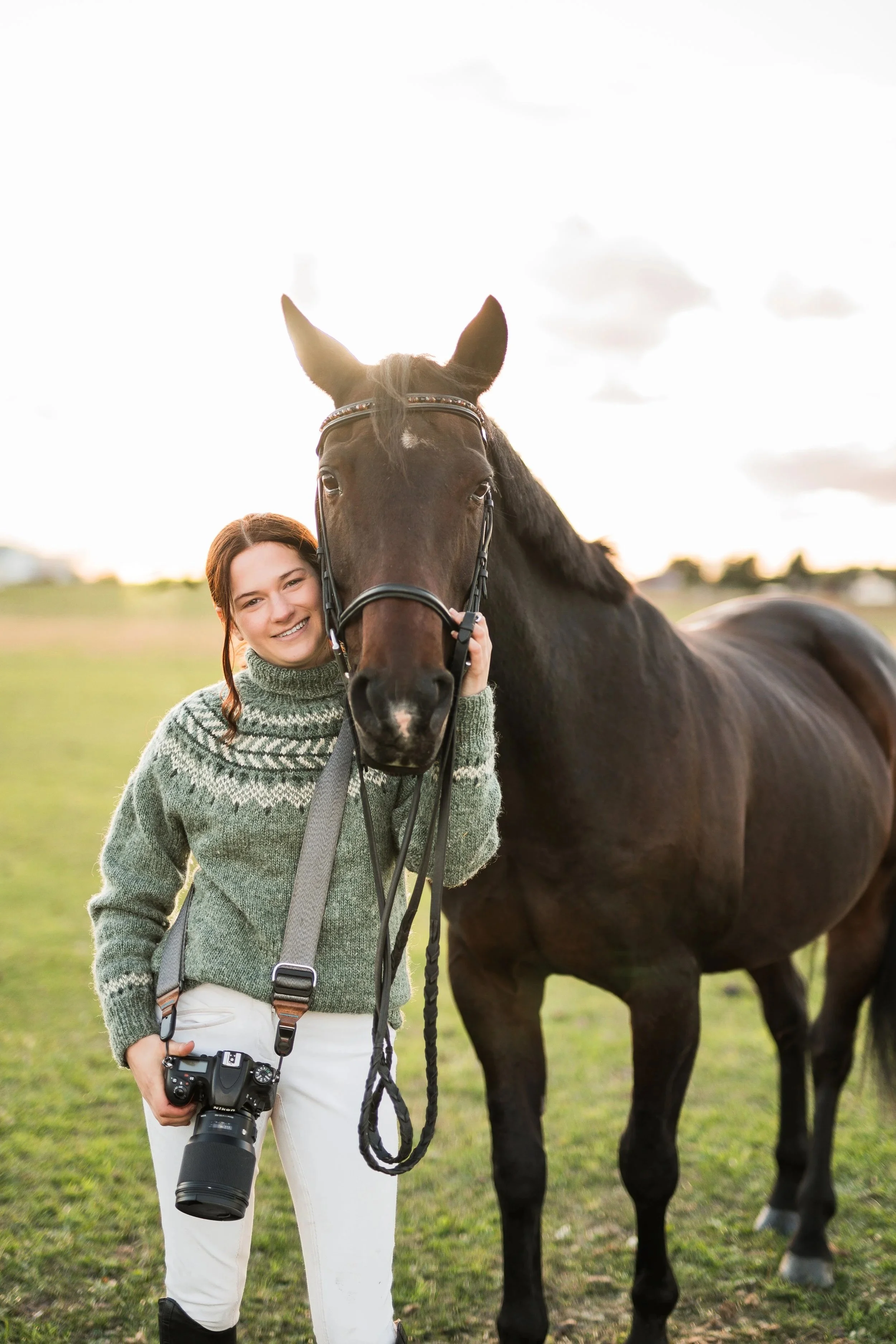 A woman with a green sweater and white pants stands outdoors holding a camera with a large lens and a horse halter in her hand. A brown horse with a bridle stands beside her. The background shows a grassy field and a cloudy sky during sunset or sunrise.