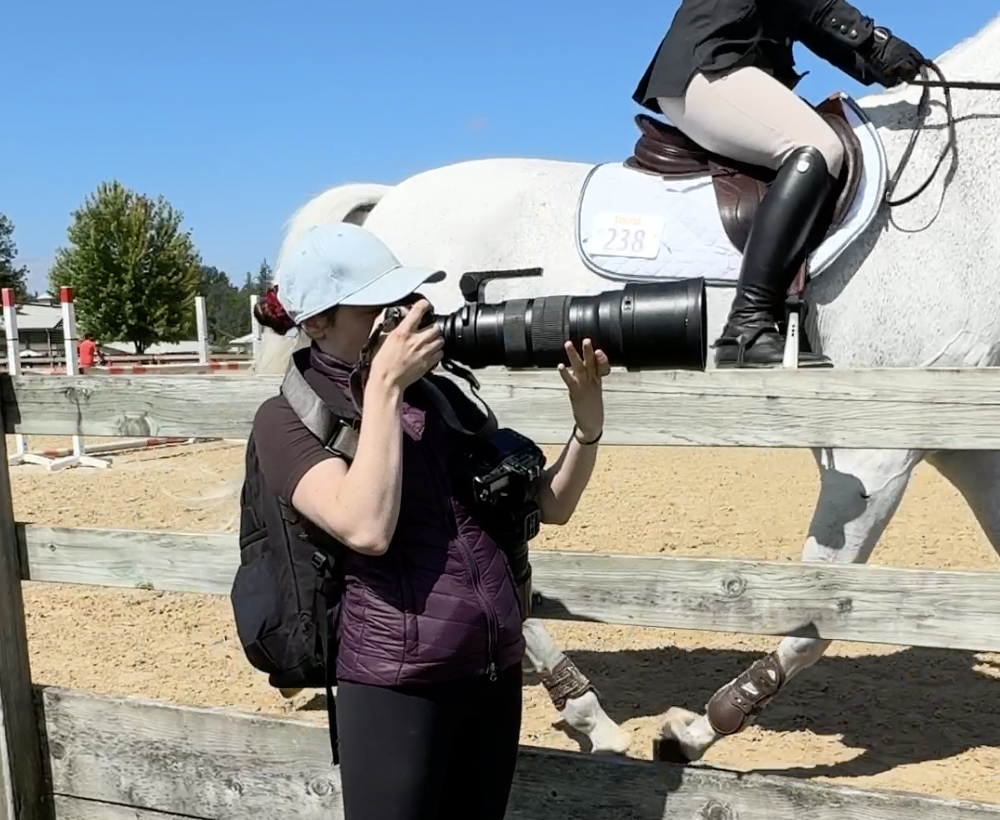 A woman taking a photograph at an equestrian event while a rider on a white horse jumps over a fence in the background.