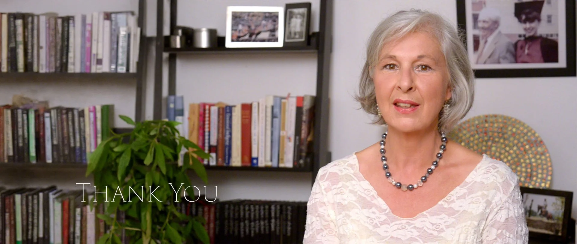 An elderly woman with short gray hair, wearing a white lace top and a pearl necklace, sitting in a room with bookshelves, framed photos, and a painting behind her. The words "Thank You" are displayed in the lower left corner.