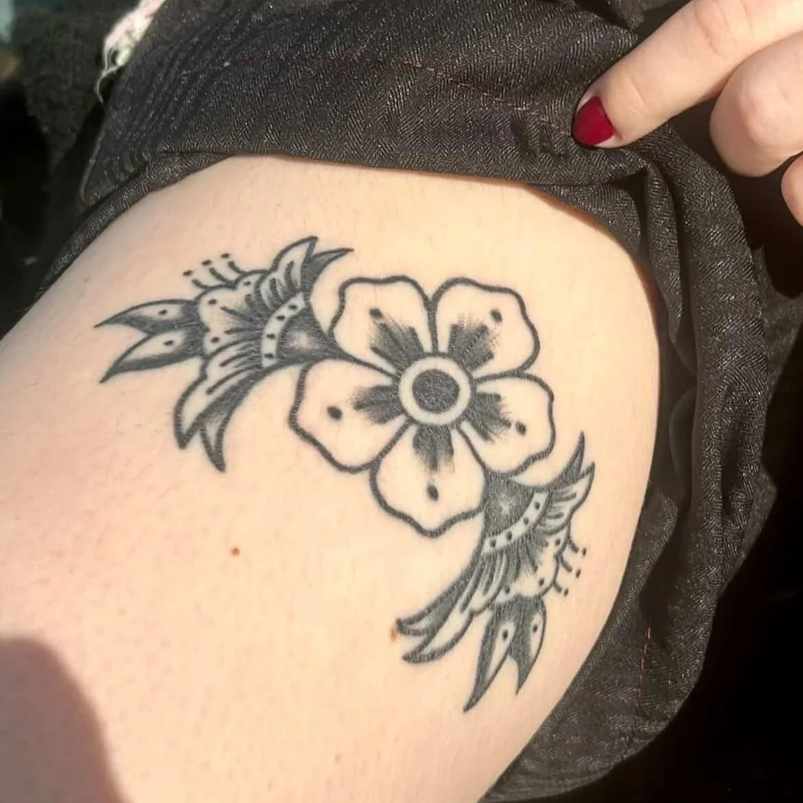 Black and gray floral tattoo on a person's thigh, featuring a large flower with petals and leaves.