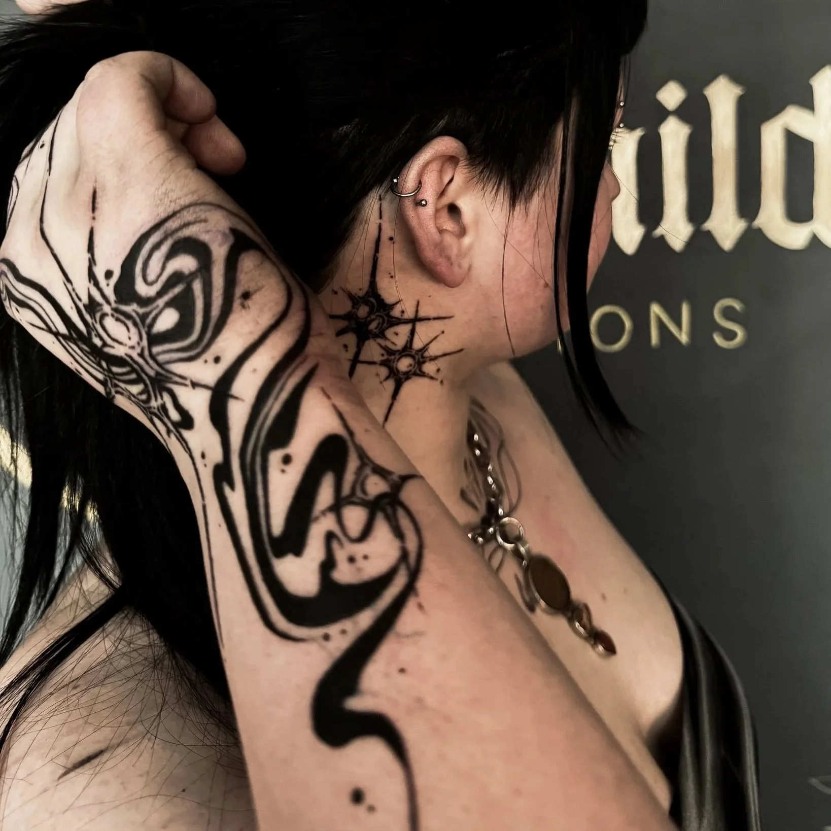 A woman with black hair and multiple tattoos, including a large tattoo of a face on her upper arm, a spiderweb with stars on her neck, and a chain necklace, has her hand behind her neck.