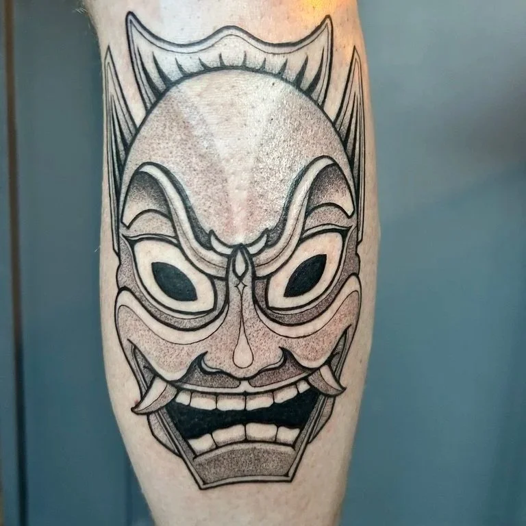 Black tattoo of a traditional Japanese oni mask on a person's thigh, featuring a fierce expression, large eyes, and prominent horns.