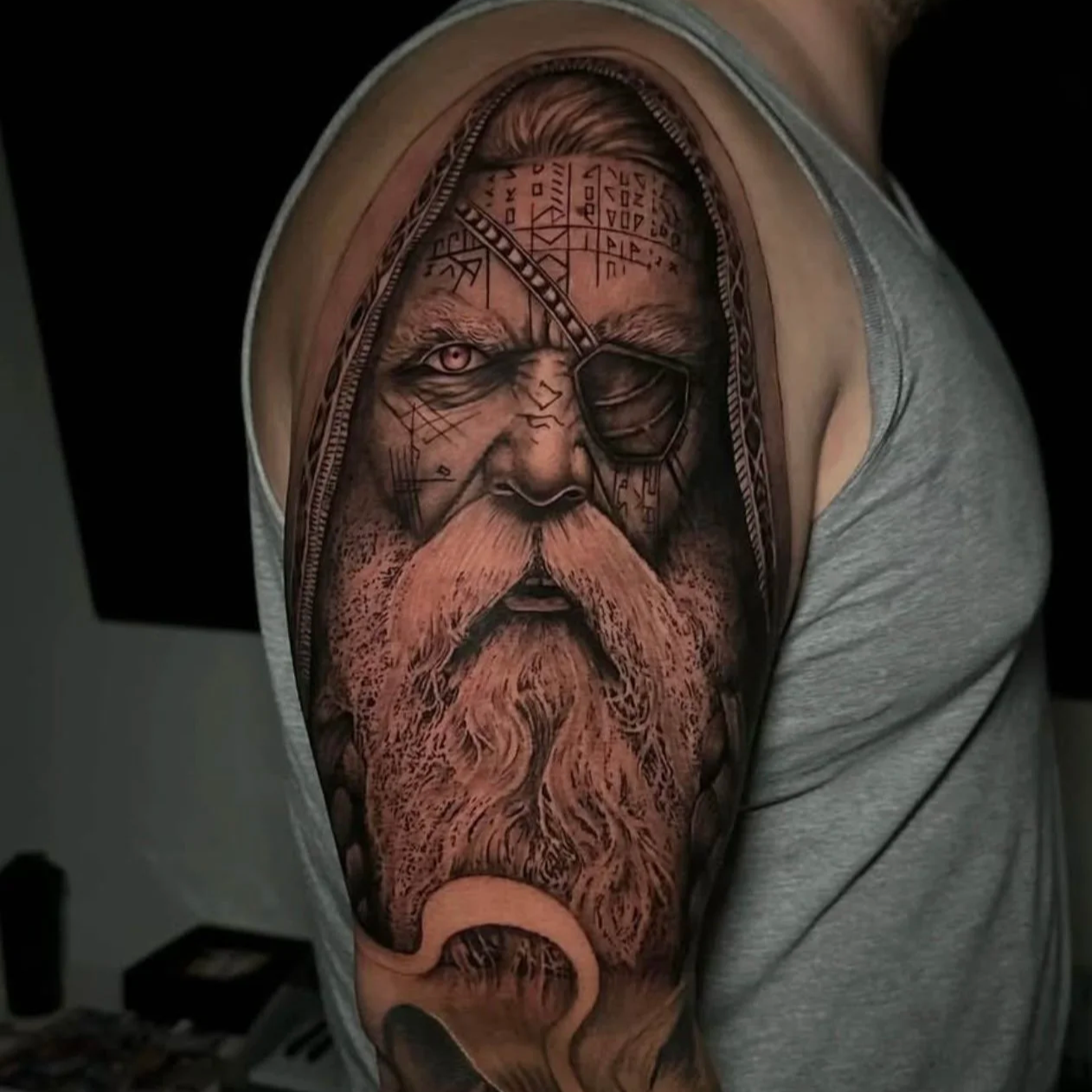 Tattoo of an old man with a long beard, eye patch over one eye, and detailed facial markings, on a person's upper arm.