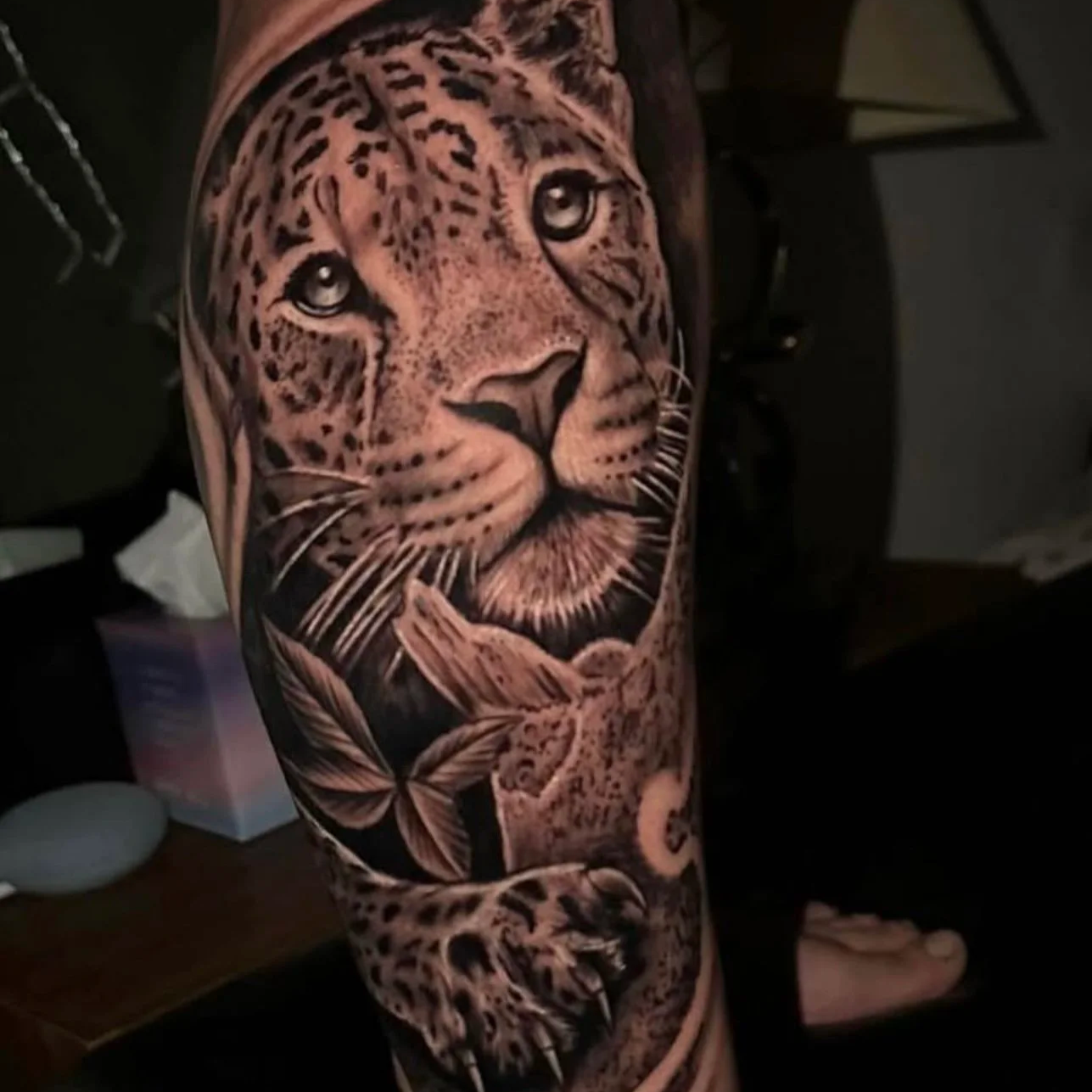 Black and gray realistic tattoo of a leopard's face on a person's arm, with detailed fur, eyes, and surrounding leaves.