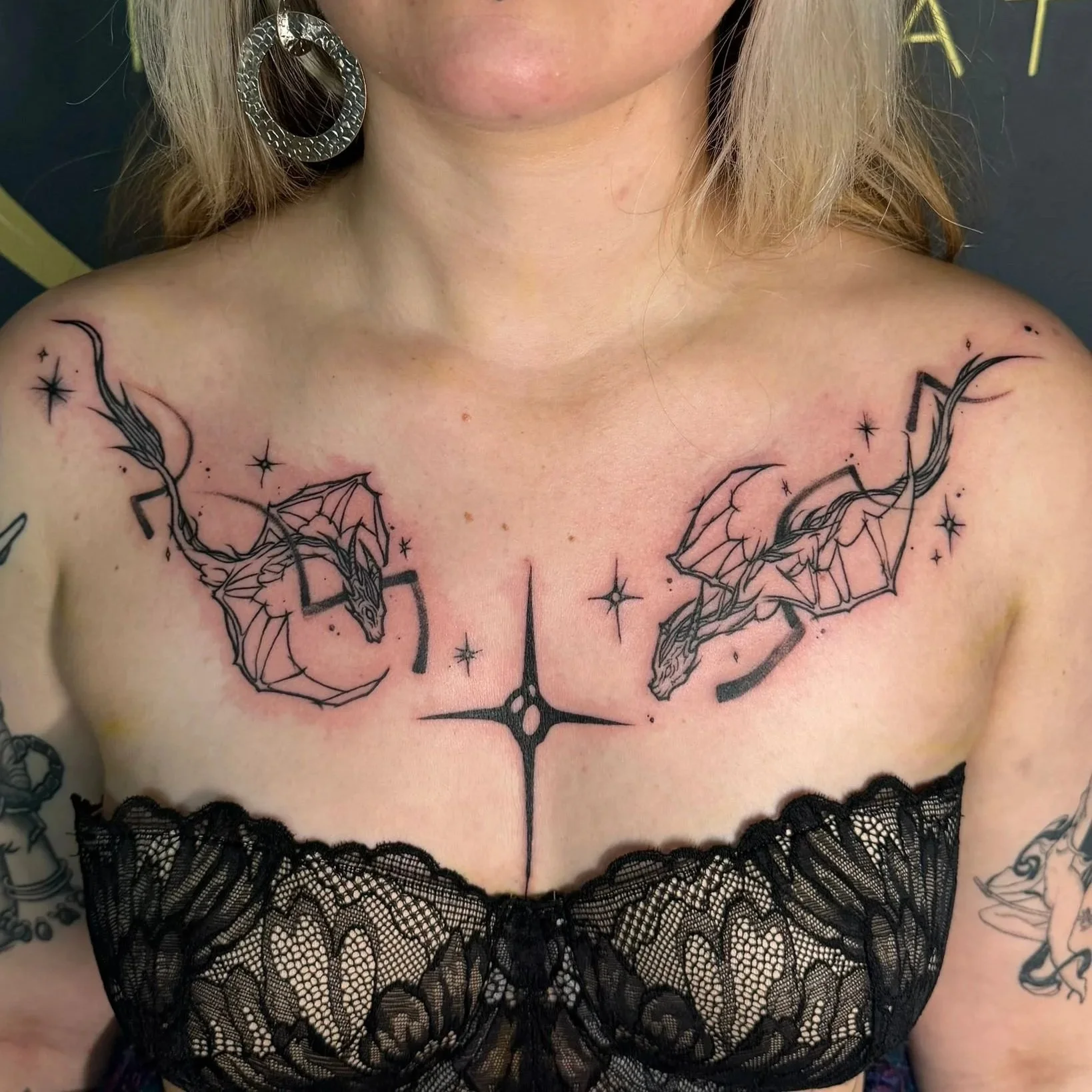 Tattoo of two dragons facing each other on the chest, with a star-shaped symbol between them, surrounded by small stars and sparkles.