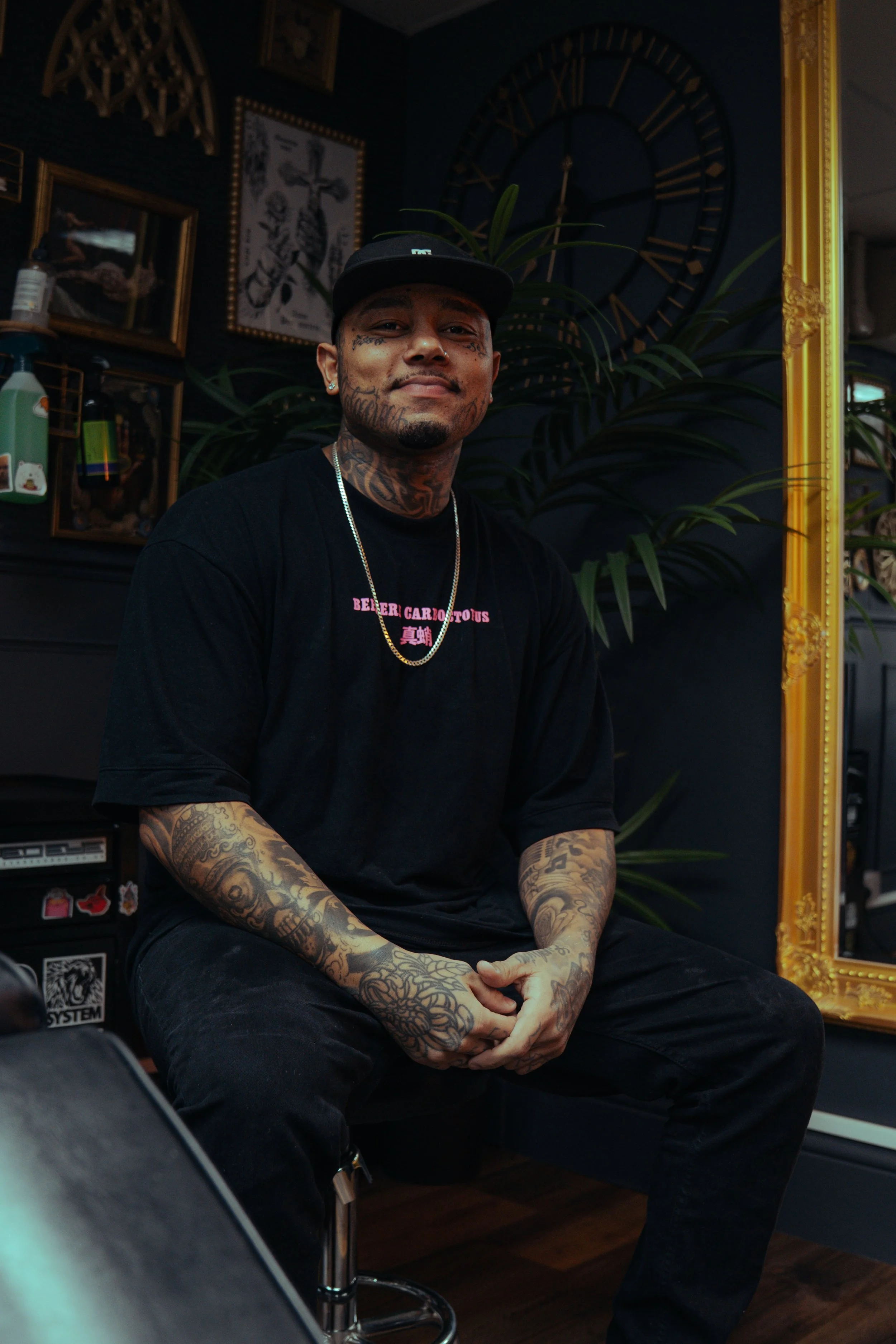 A man with tattoos sits on a stool in a dimly lit room, with a large clock and various framed pictures on the dark wall behind him. He wears a black cap, black t-shirt, and jewelry, with a relaxed expression.