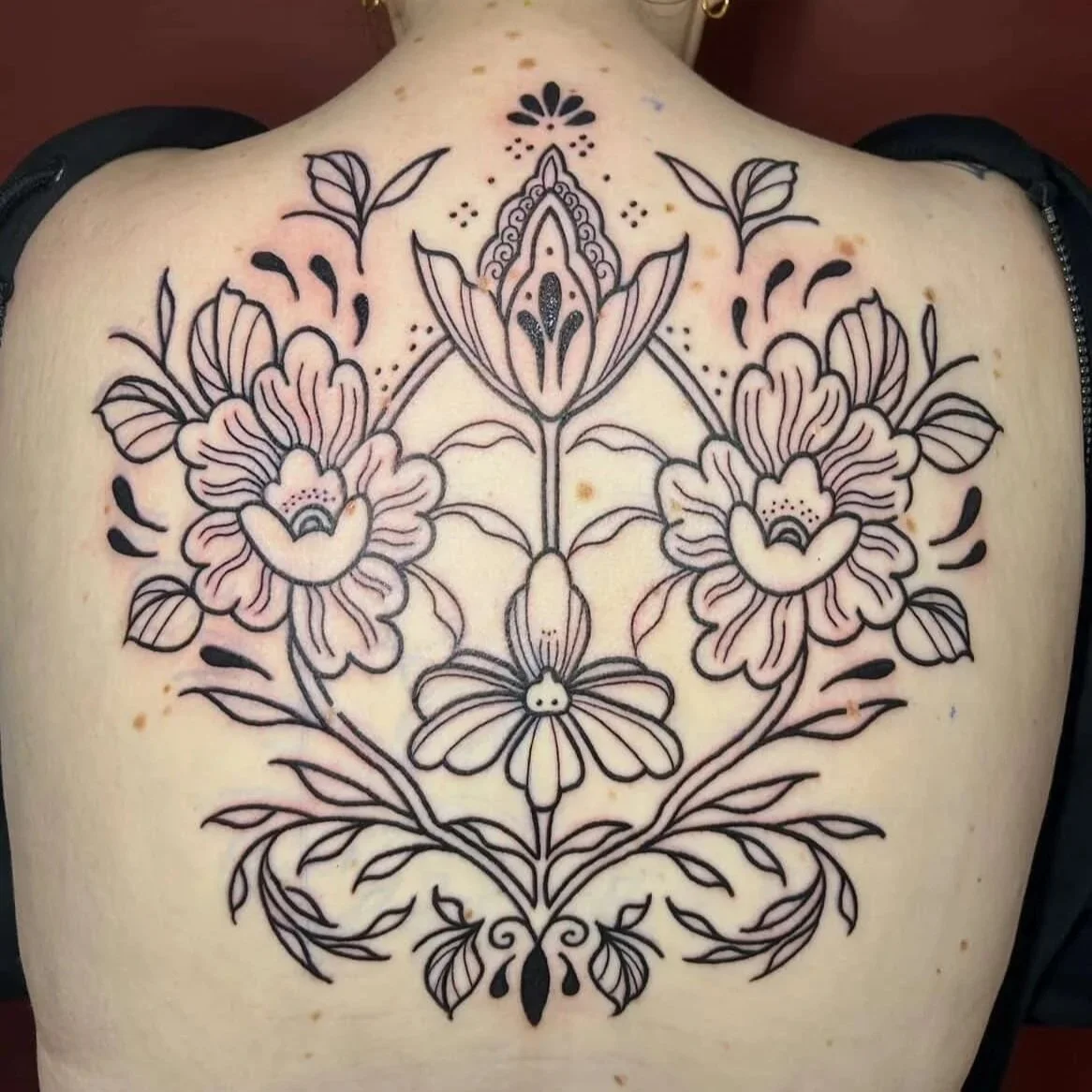 A black outline floral tattoo on person's back featuring large flowers, leaves, and symmetrical decorative elements.