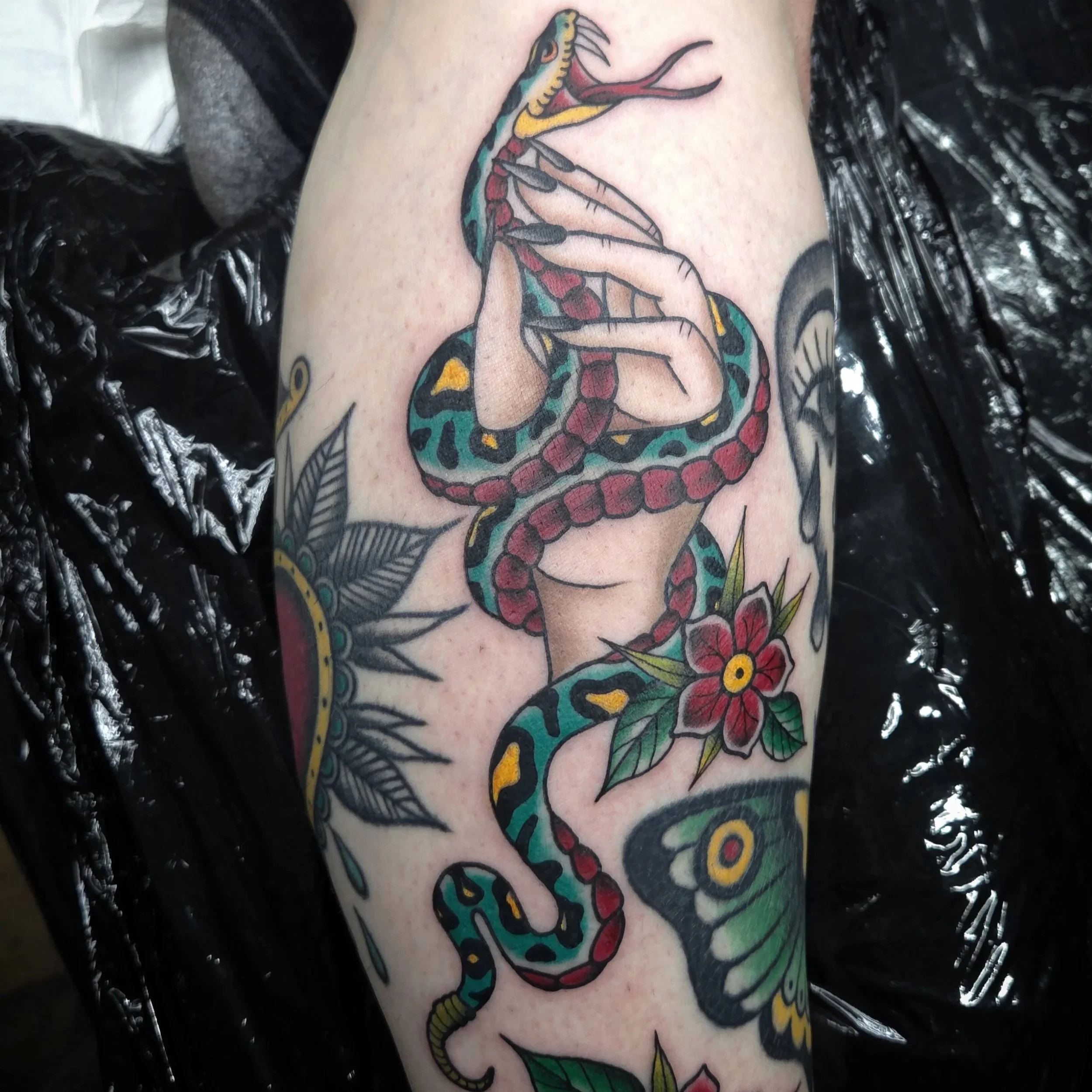Tattoo of a hand holding a colorful snake, surrounded by flowers, leaves, a butterfly, and abstract designs.