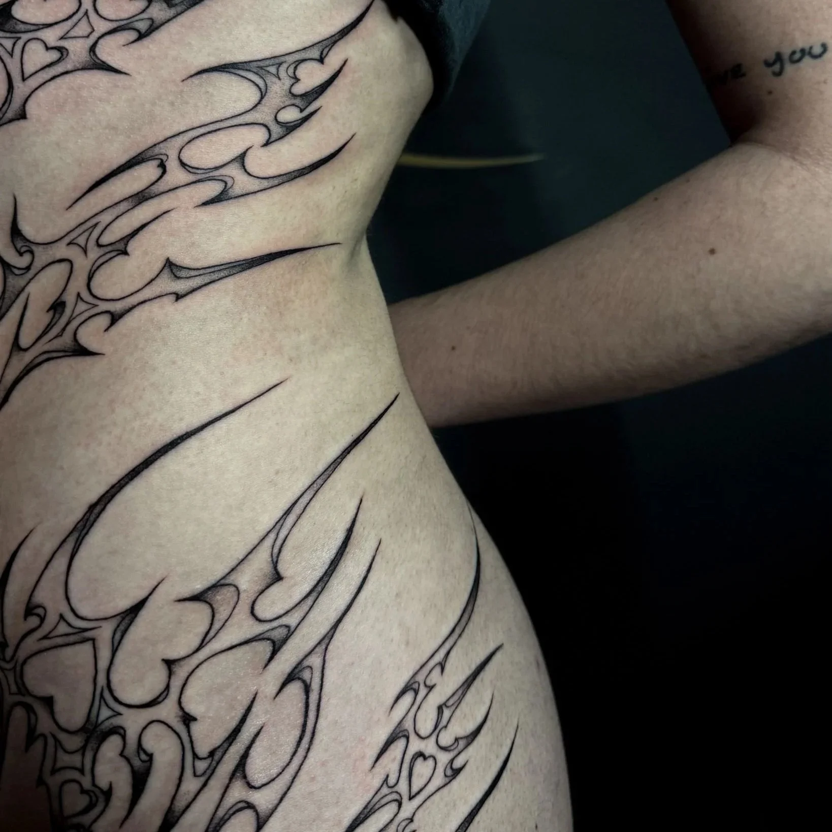 Close-up of a person's side and torso with detailed black tribal tattoos featuring sharp, flowing lines and heart shapes.