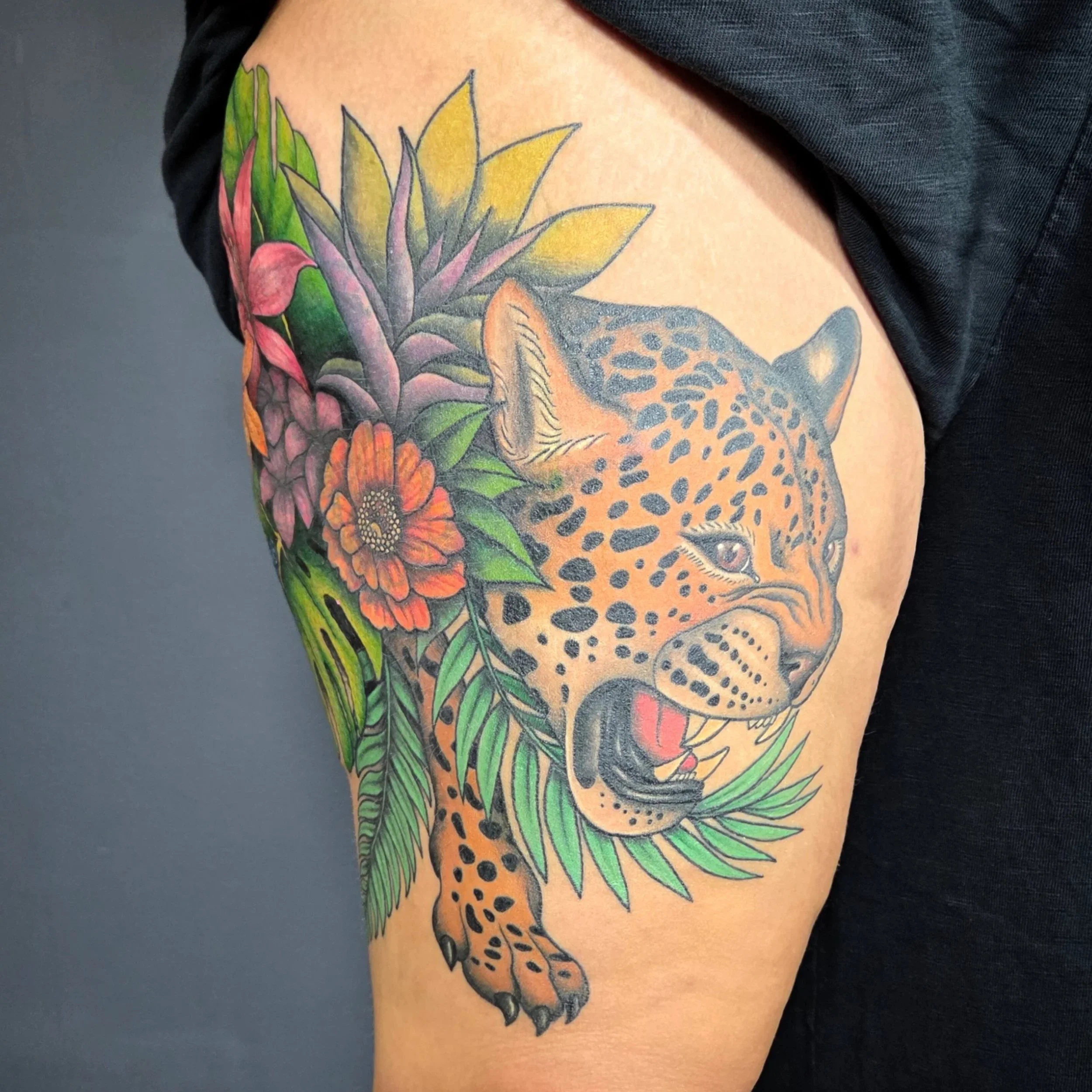 Colorful tattoo of a jaguar's face surrounded by tropical flowers and leaves on a person's upper arm.