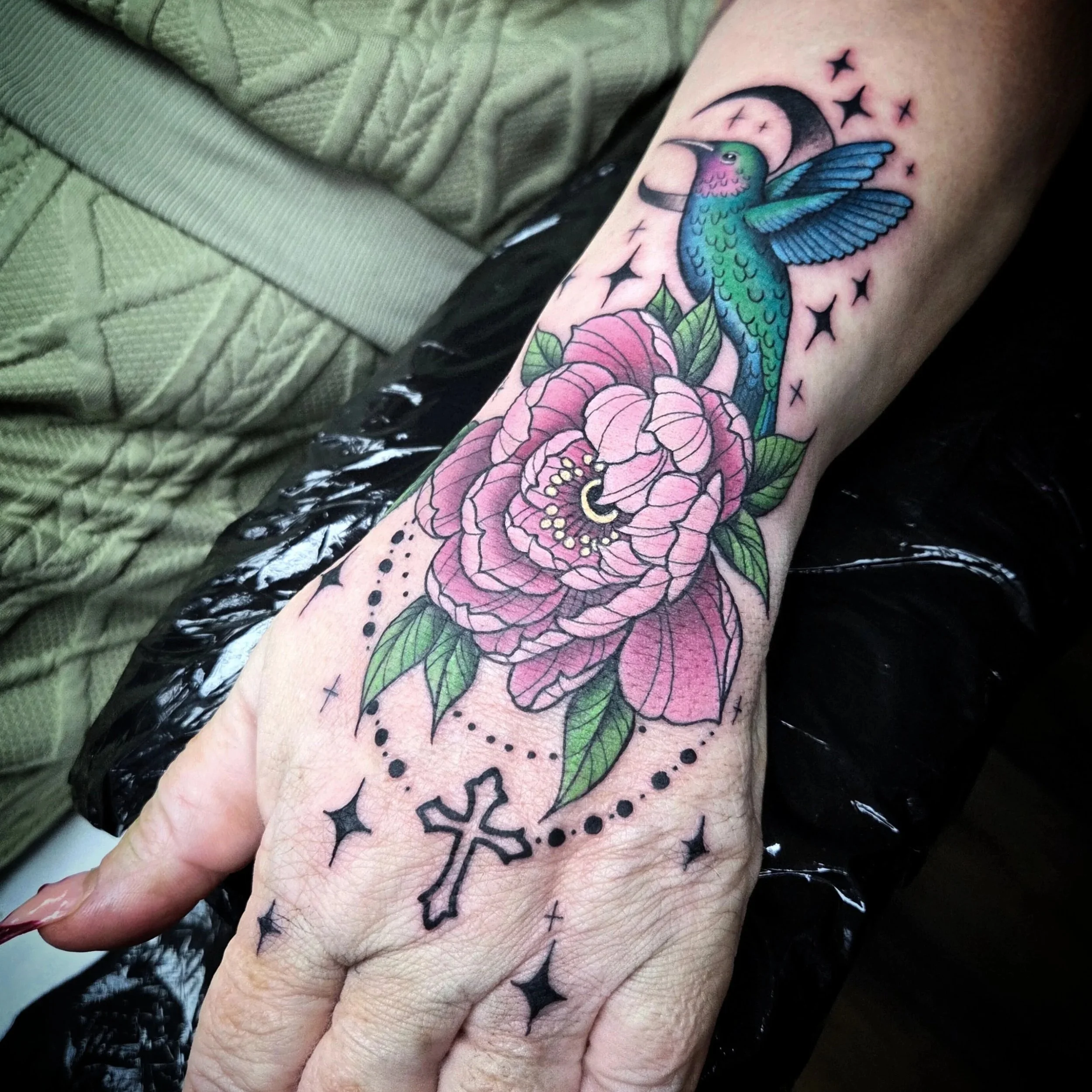 Tattoo of a hummingbird and pink flower on a person's forearm, with stars, dots, and a cross surrounding the design.
