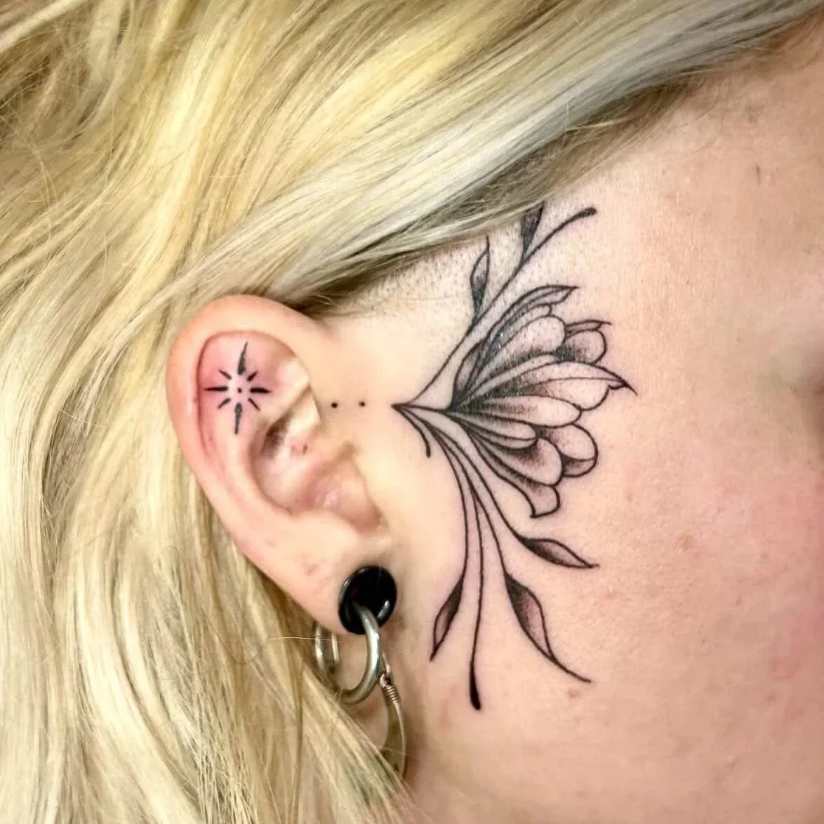 Close-up of a person's ear with multiple tattoos, including a butterfly or flower design behind the ear, and a small star and dots near the ear lobe. The person has blonde hair and multiple earrings.