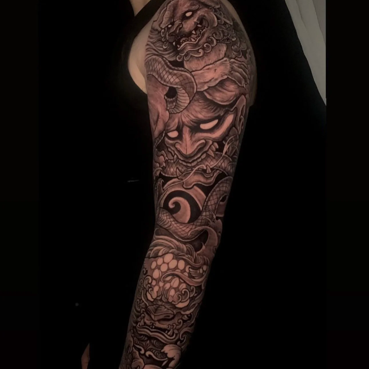 Black and gray tattoo sleeve featuring a fierce lion, a human face with an intense expression, and various swirling cloud and dragon motifs.