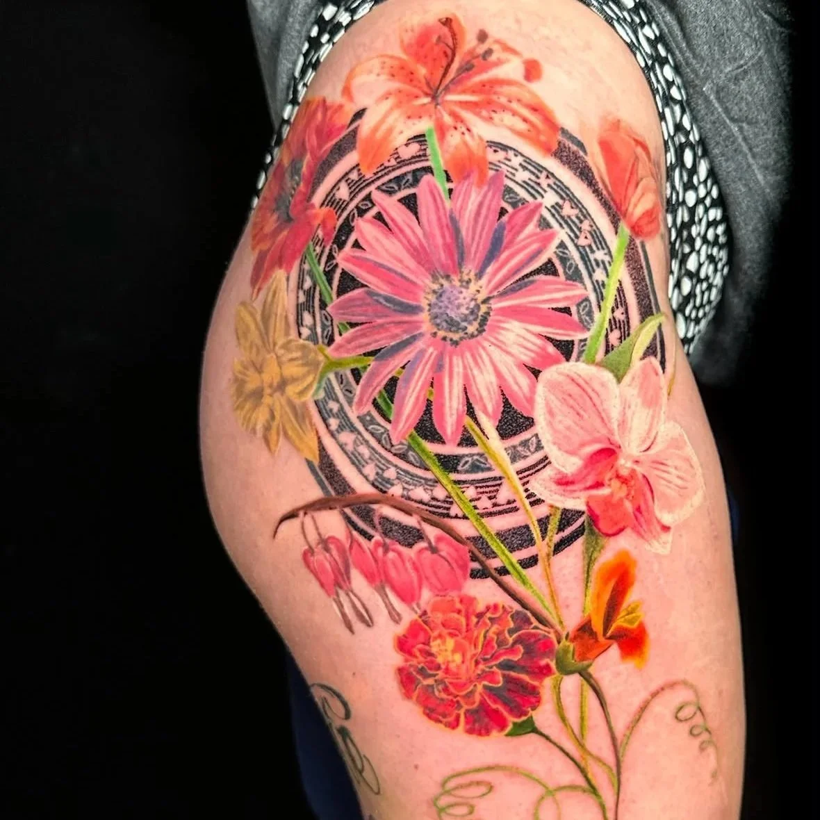 Colorful tattoo of various flowers including lilies, daisies, orchids, and carnations with a mandala design in the background on a person's upper arm.