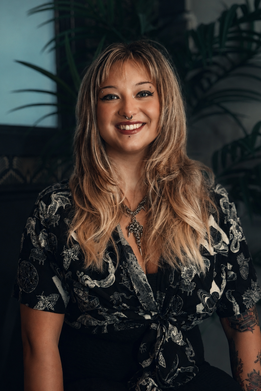 A smiling woman with blonde, wavy hair and piercings on her ears and nose, wearing a black shirt with white designs and a necklace, sitting in front of a dark background with green plants.