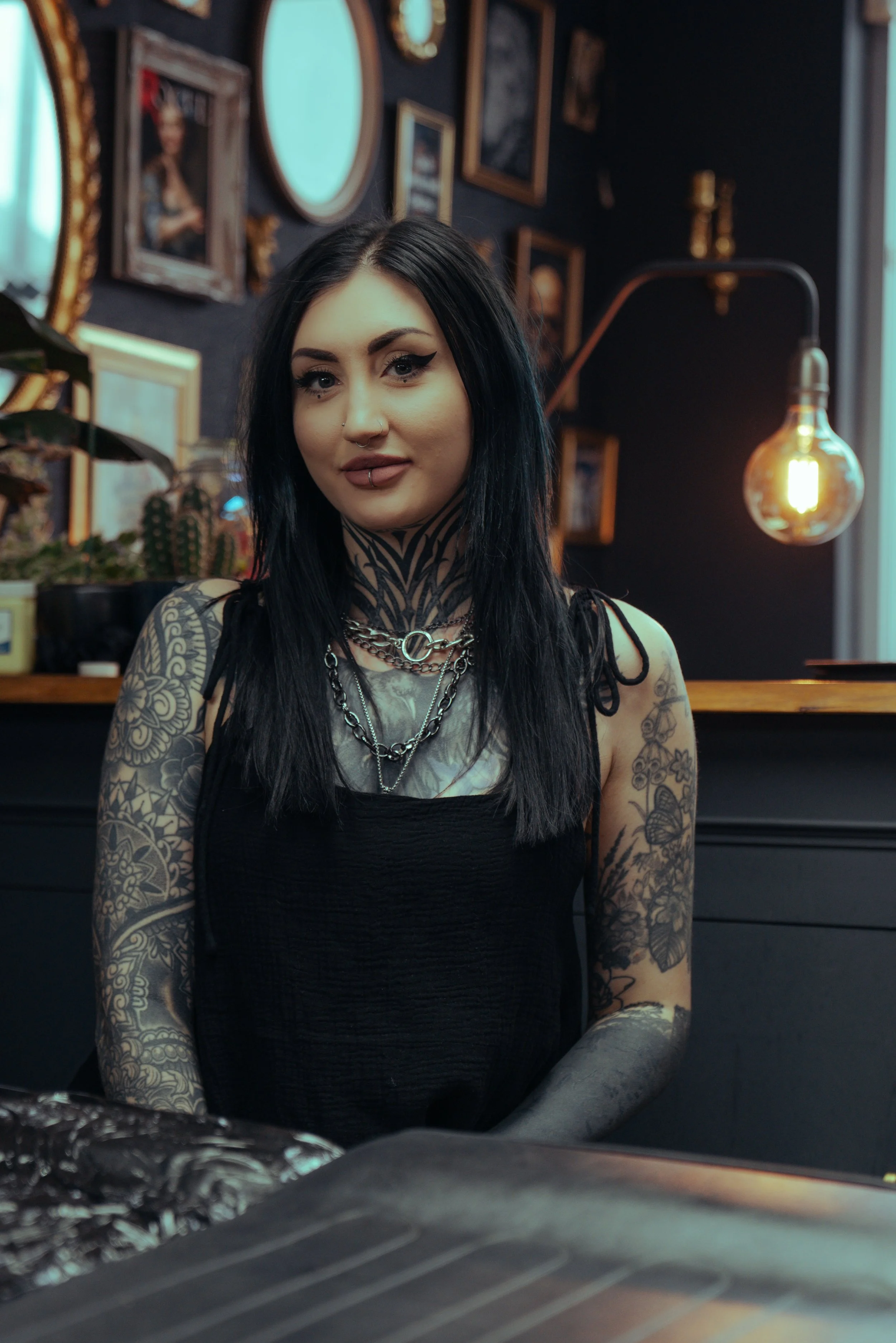 A young woman with black hair and tattoos on her arms and neck, wearing a black top and jewelry, standing inside a room with dark walls decorated with framed pictures and mirrors, and a hanging lightbulb.