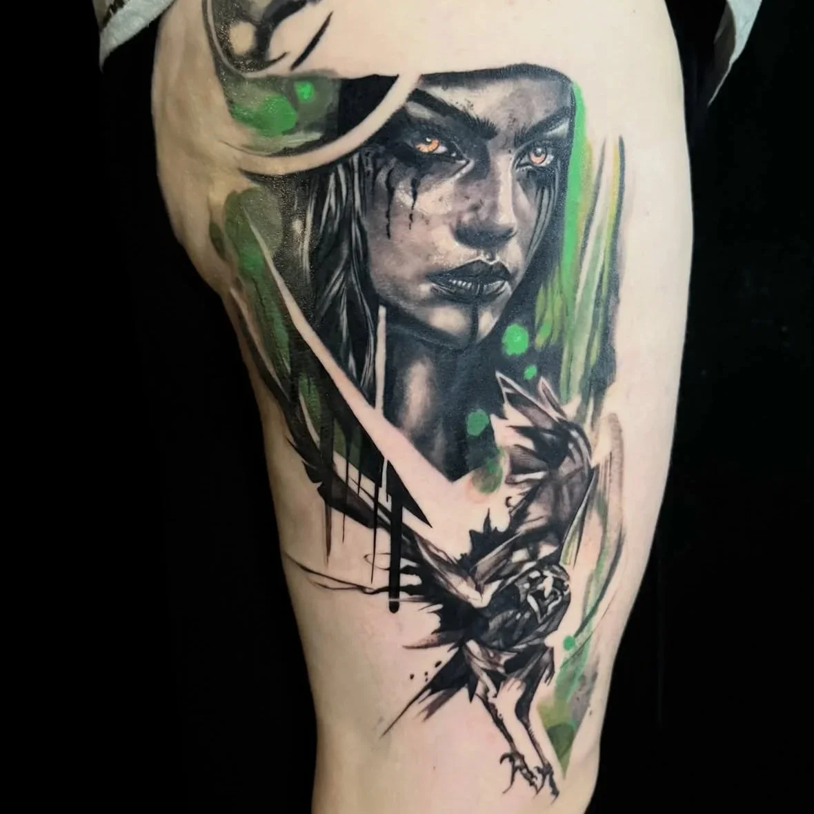 Tattoo of a woman's face with dark makeup, long hair, and intense eyes, surrounded by abstract green, black, and pink artistic elements.