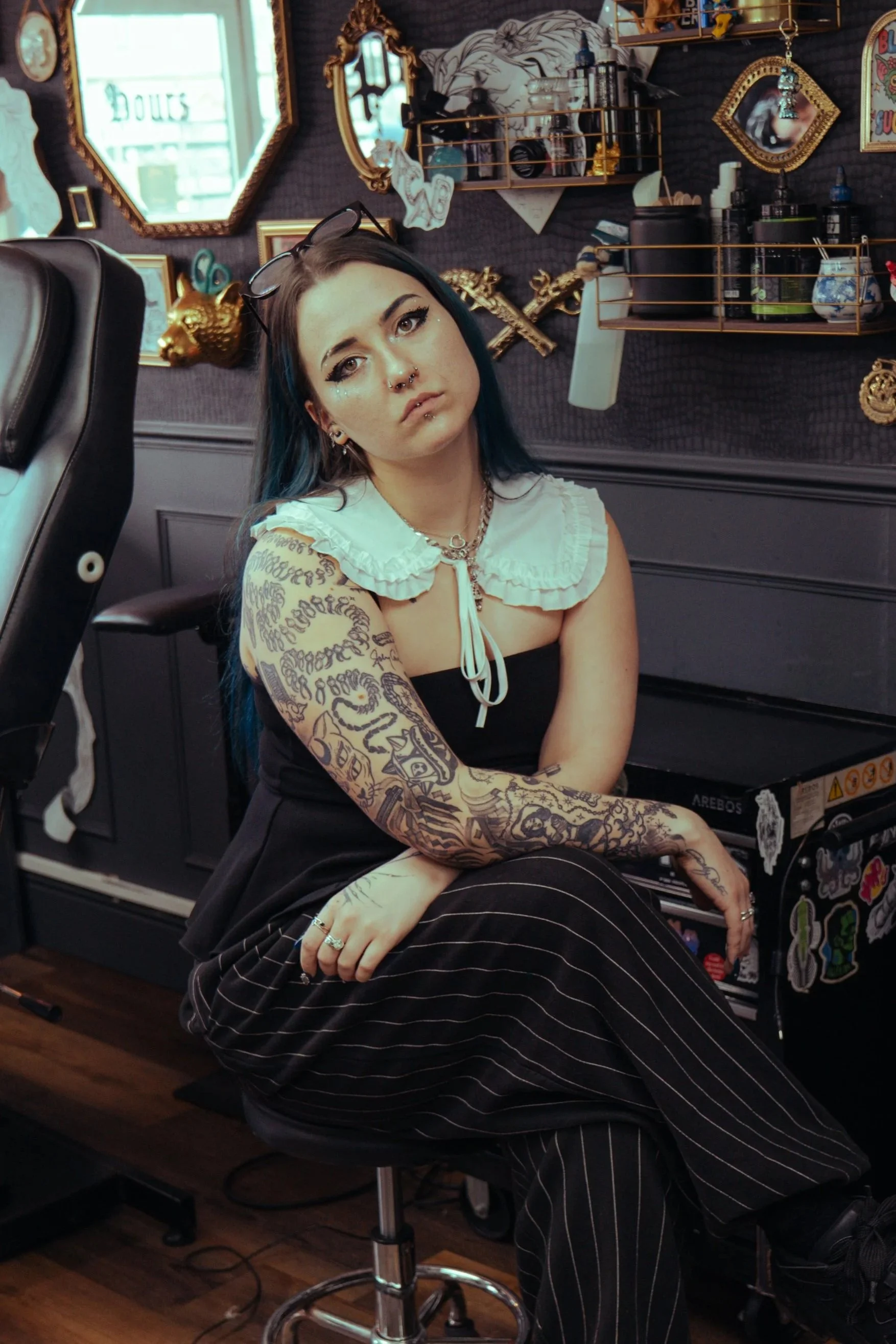 A young woman with blue and black hair, tattoos on her arm, and multiple piercings on her face, sitting in a tattoo studio with a dark wall decorated with mirrors, shelves, and framed artwork.