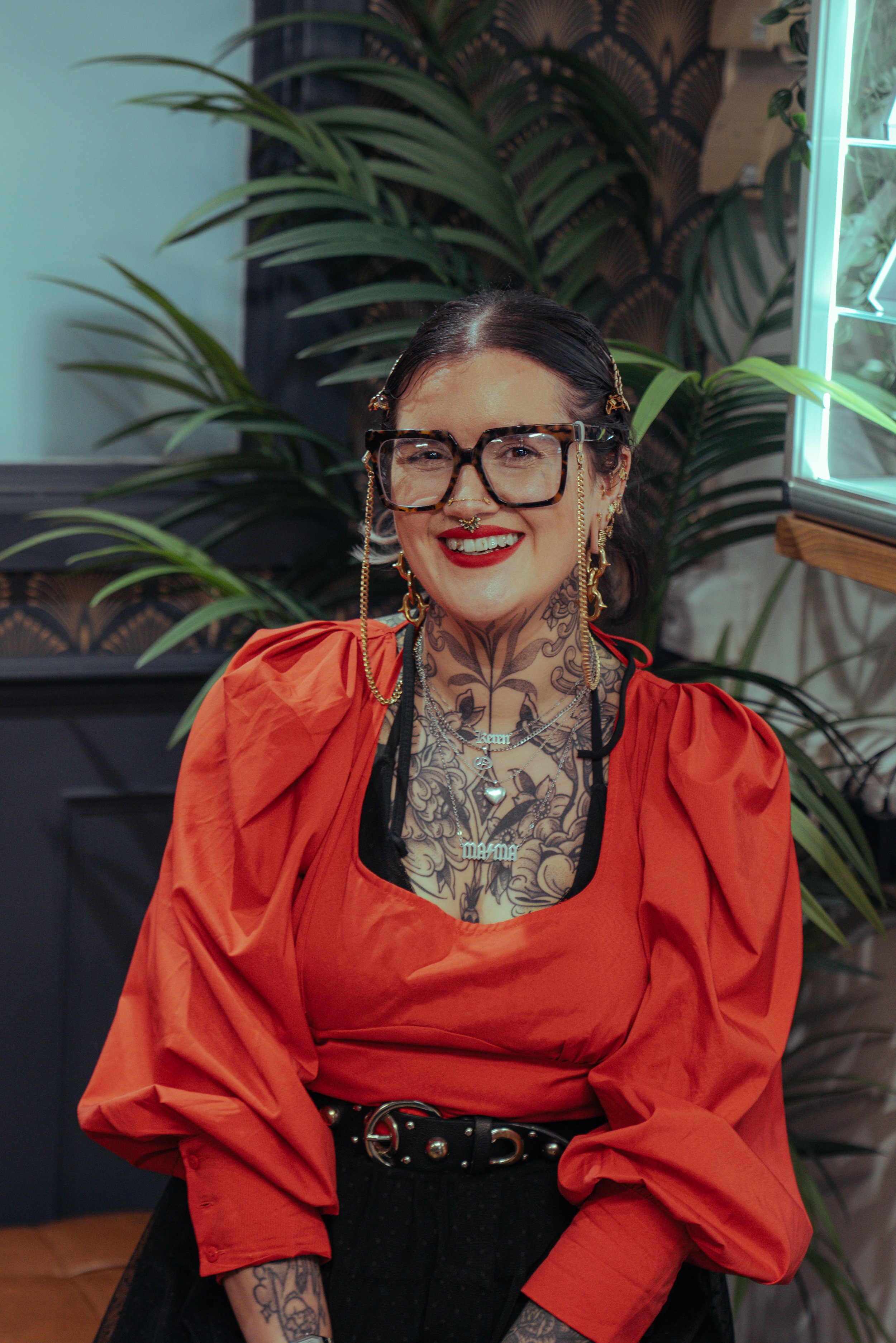 A woman with dark hair, tattoos, and large two-toned glasses smiling and wearing an orange top with a black belt, standing in front of a leafy background with a neon light on her right.