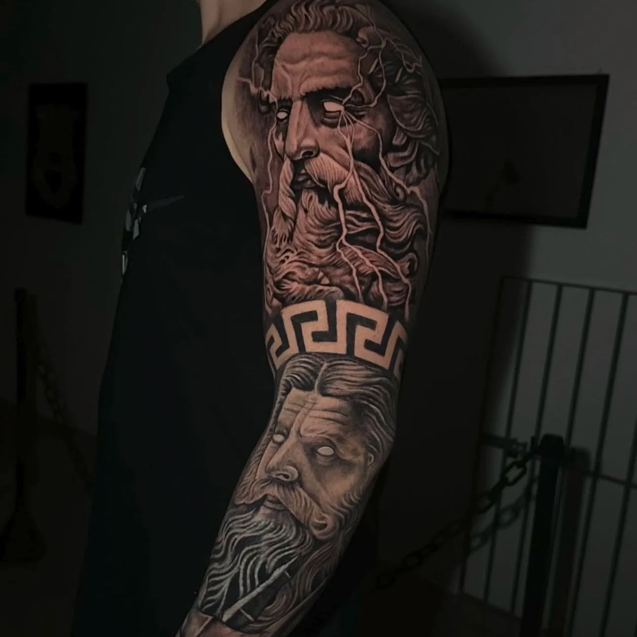Close-up of a person's arm showing a detailed black and gray tattoo sleeve featuring a classical bearded philosopher with flowing hair and beard, and a Greek design band.