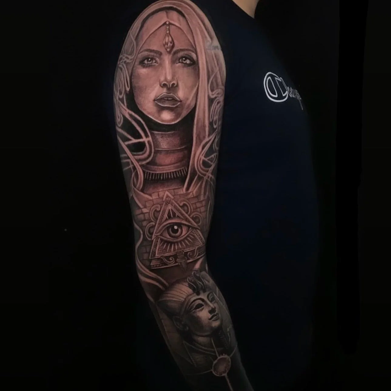Tattoo sleeve on a person's upper arm featuring a realistic portrait of a woman with long hair, a prominent eye within a pyramid below her, and other decorative elements in black and gray ink.
