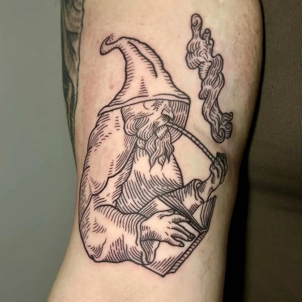 Tattoo of a wizard with a hat, beard, and pipe, holding an open book, with smoke curling upward.