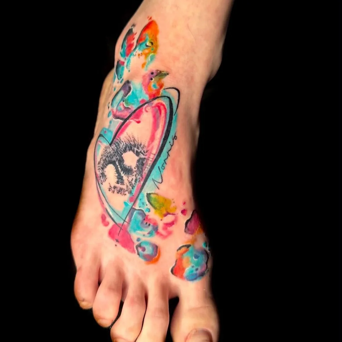 Colorful watercolor-style tattoo on a foot, featuring a heart shape with a skull inside and abstract shapes and splashes in various bright colors.