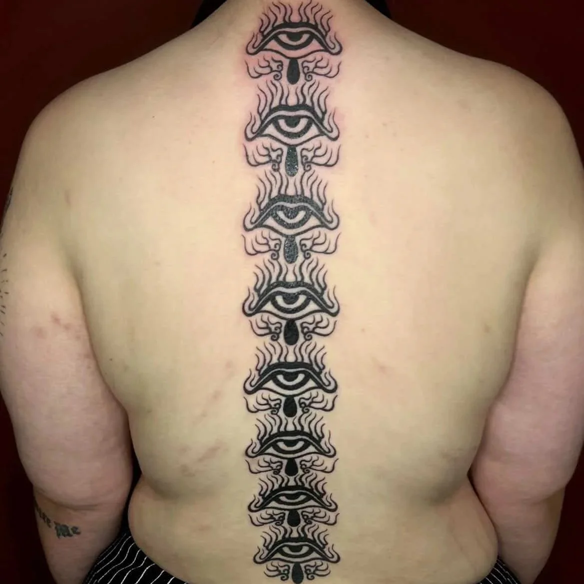Tattoo design of multiple eyes with teardrops and flames running down the center of a person's back.
