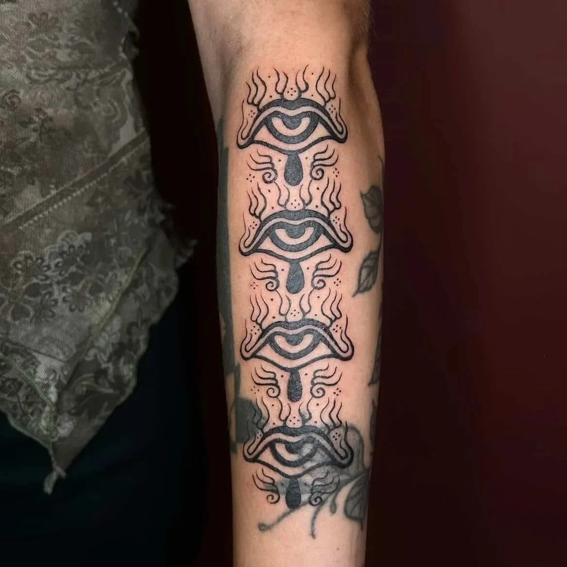 Tattoo of stylized eyes with flames and teardrops, repeated vertically along the forearm.