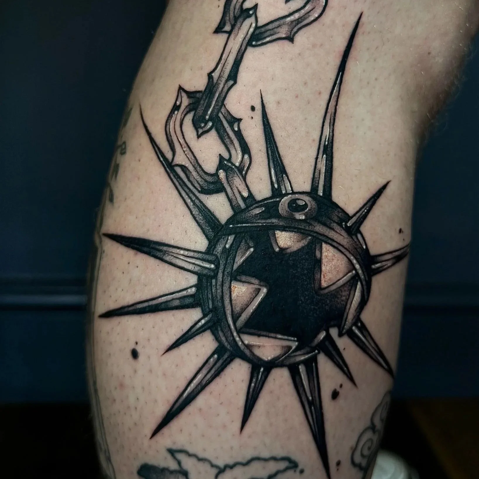 A black ink tattoo of a star with sharp, elongated points and an eye at the center, surrounded by additional lines and details.