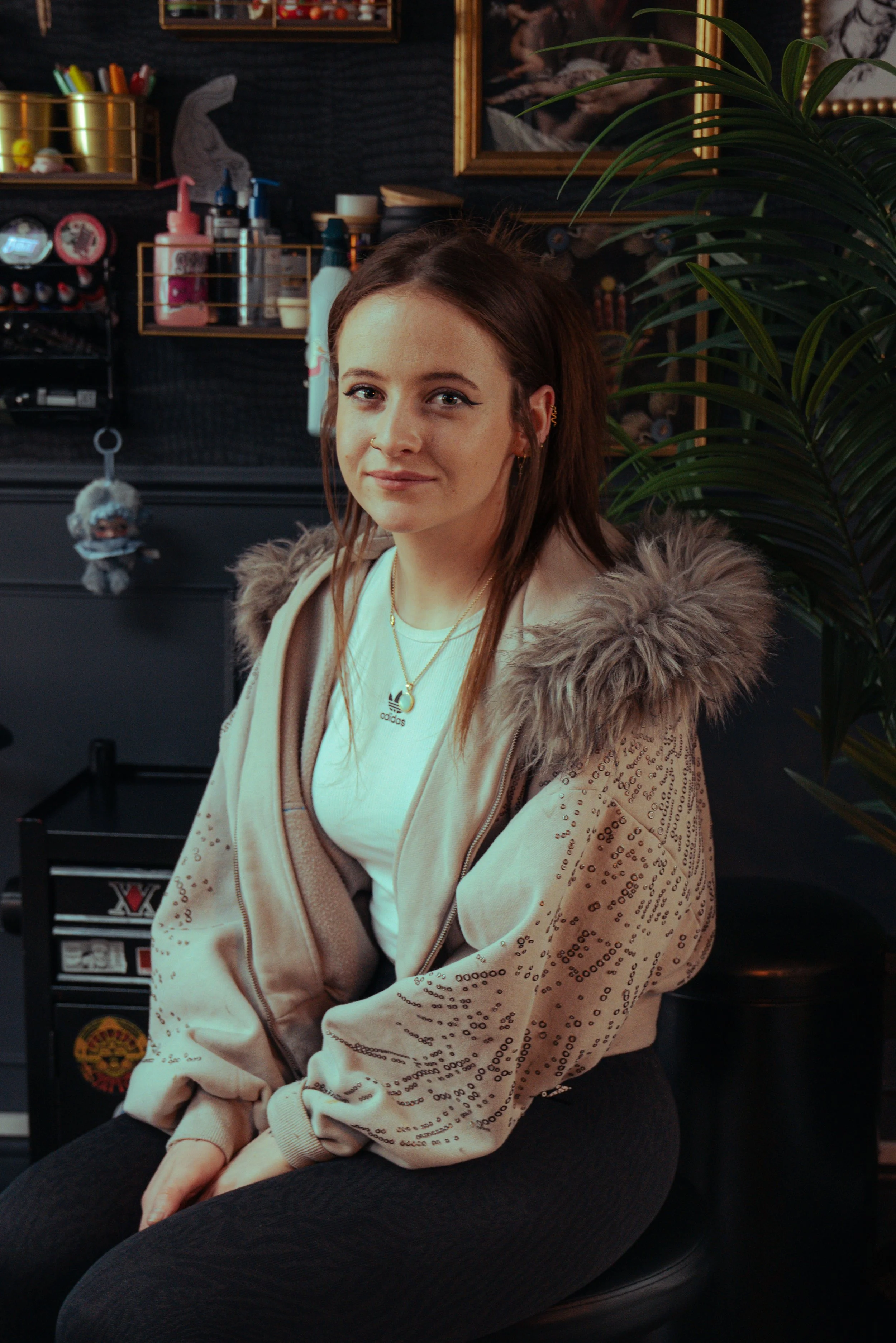 A young woman with brown hair, wearing a beige zip-up hoodie with a fur-lined hood, a white Adidas t-shirt, and black pants, sitting with her hands on her lap, in a room with dark walls, a large houseplant, and shelves with miscellaneous items.