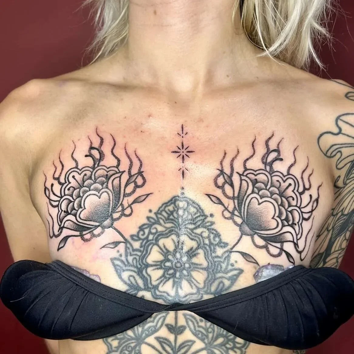 A woman's chest with multiple black and gray tattoos including flowers, a symmetrical mandala, and decorative elements.