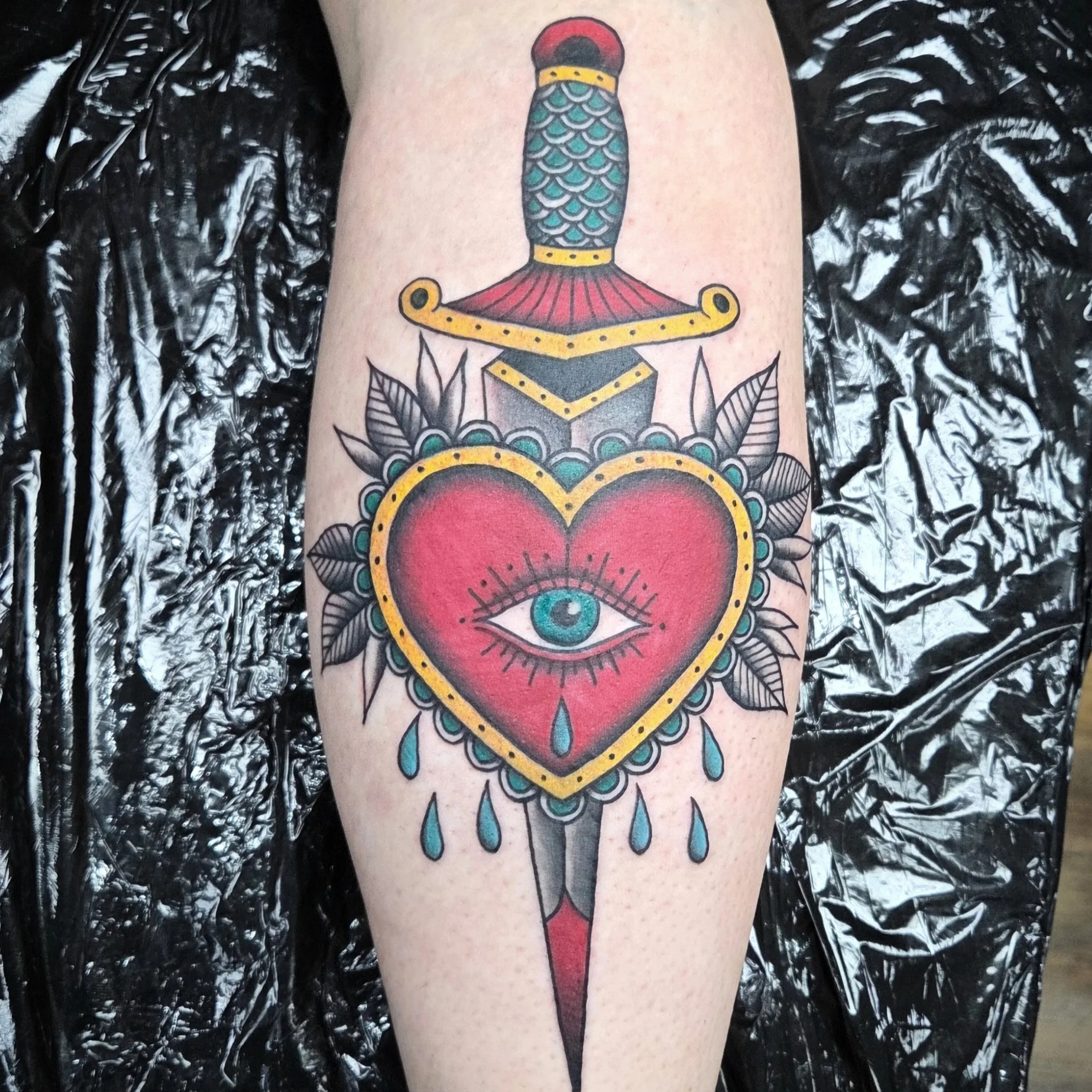 Tattoo of a sword piercing through a heart with an eye in the center, surrounded by leaves and blue tears.