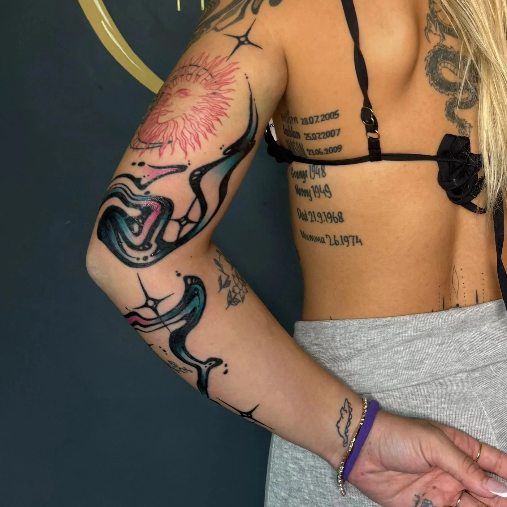 A woman with tattoos on her arm and torso, wearing a black strappy top and gray sweatpants.