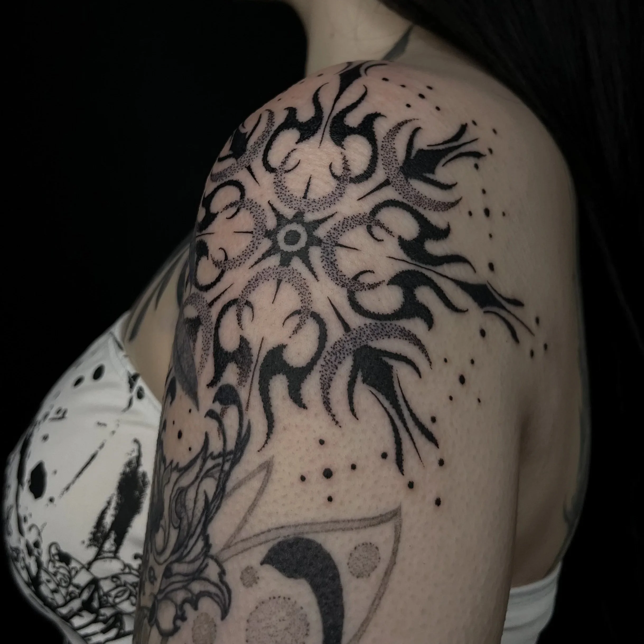 Black tribal-style tattoo on person's shoulder and upper arm featuring a star or sun design with tentacle-like extensions, dots, and abstract elements.