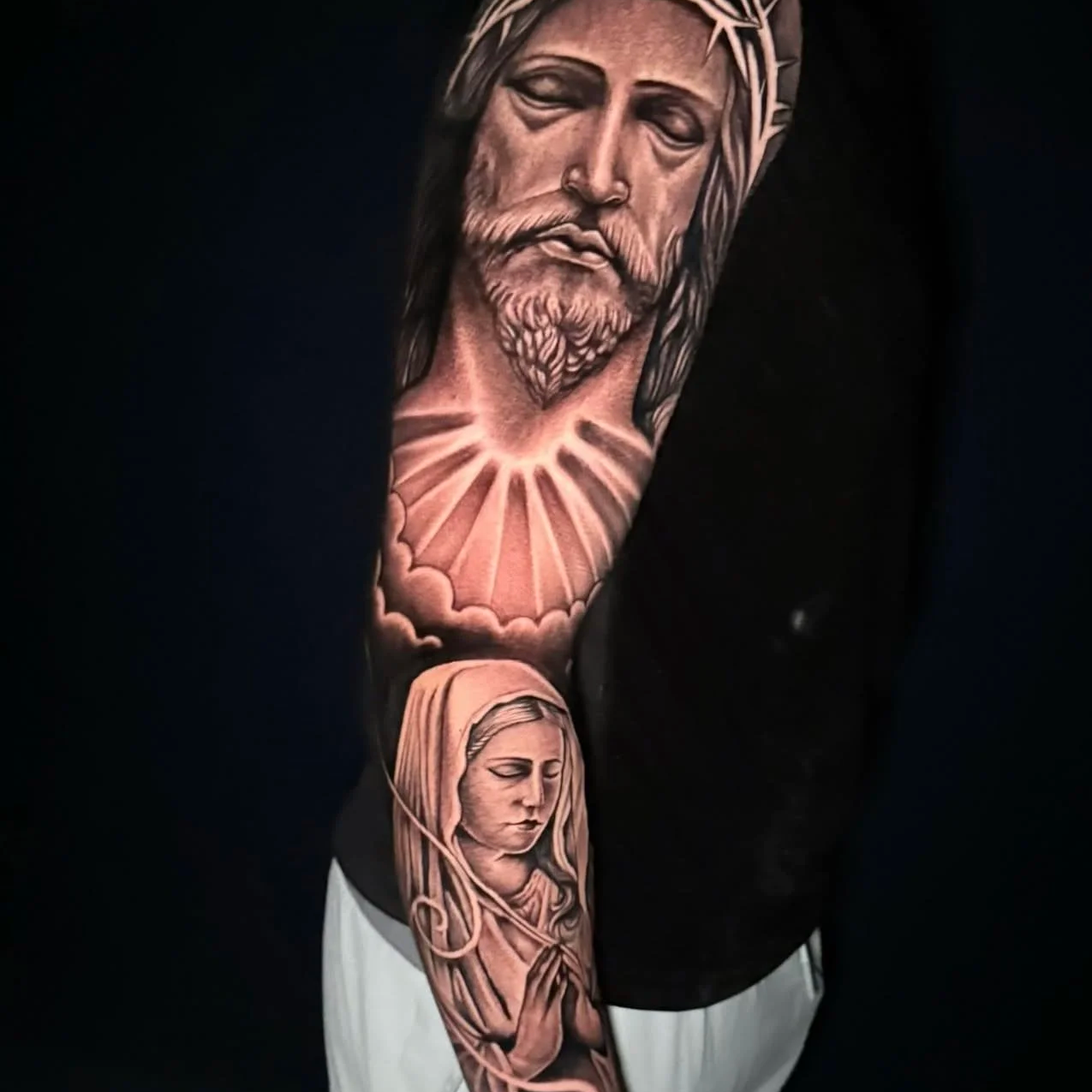 A tattoo of a Jesus Christ face with a crown of thorns, a halo of light or rays behind his head, and a praying woman with a halo and prayer beads on her arm.