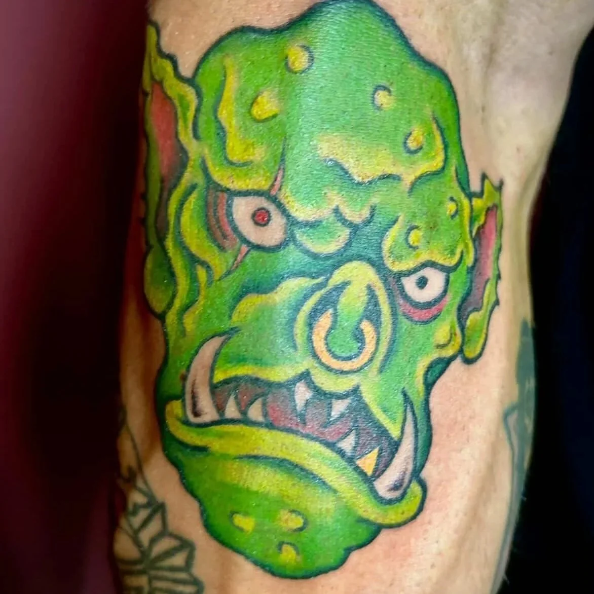 Colorful tattoo of a zombie-like face with green skin, bulging eyes, sharp teeth, and a long tongue.