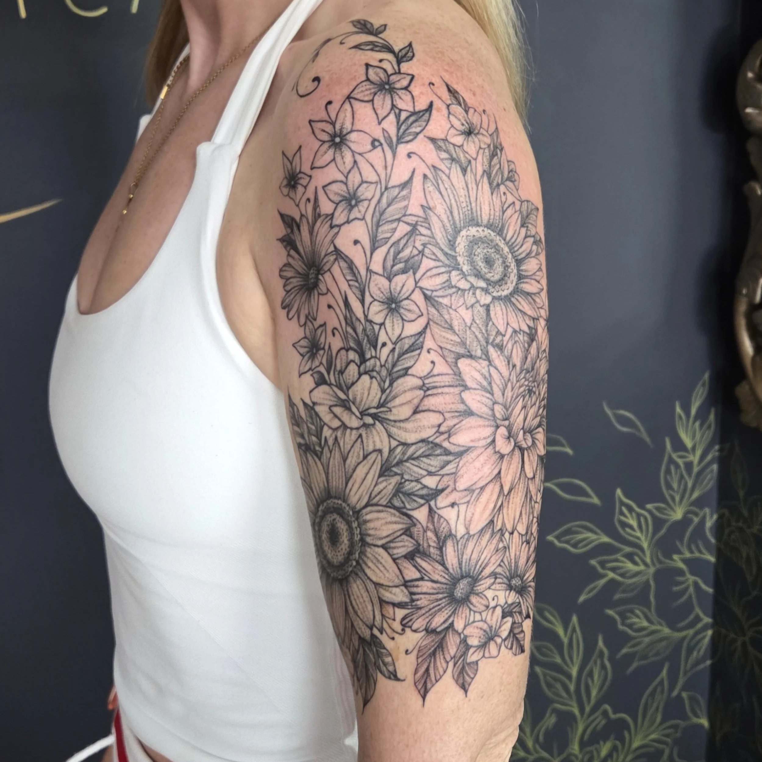 Black and white floral tattoo on woman's upper arm and shoulder, featuring sunflowers, daisies, and other flowers with detailed shading.