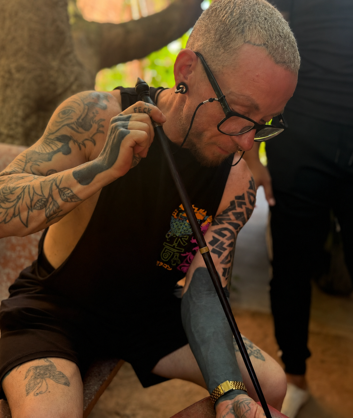 A young man with tattoos, glasses, and bleached hair is sitting and smiling while holding a walking cane. He is wearing a black sleeveless shirt and appears to be in a relaxed, outdoor setting.