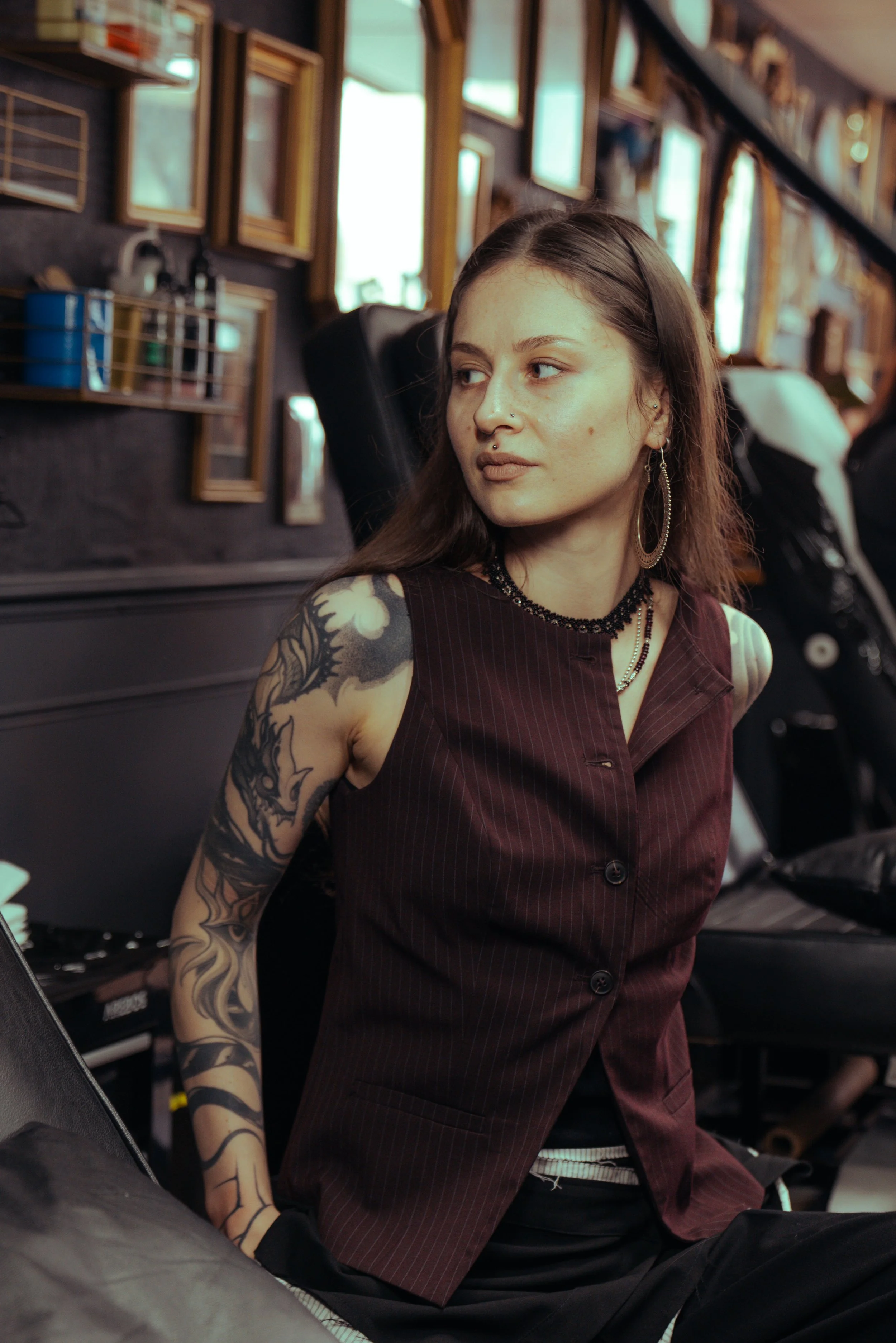 A young woman with tattoos, wearing earrings, a beaded necklace, and a dark sleeveless top, sitting in a barber shop or similar setting, with frames and mirrors on the wall behind her.