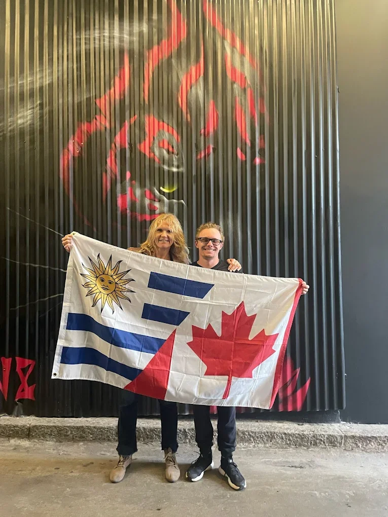 Two people standing in front of a large mural, holding a flag with the Uruguayan and Canadian flags. The mural depicts a stylized image of a roaring lion with red and black colors.