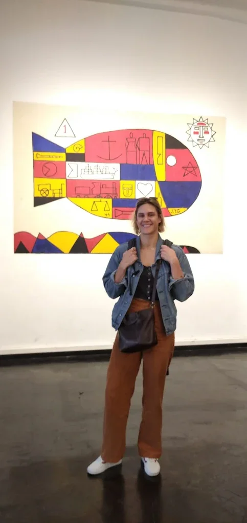 A young woman standing in front of a colorful abstract mural in an art gallery. She is smiling, wearing a denim jacket, black top, brown pants, white sneakers, and carrying a black shoulder bag.