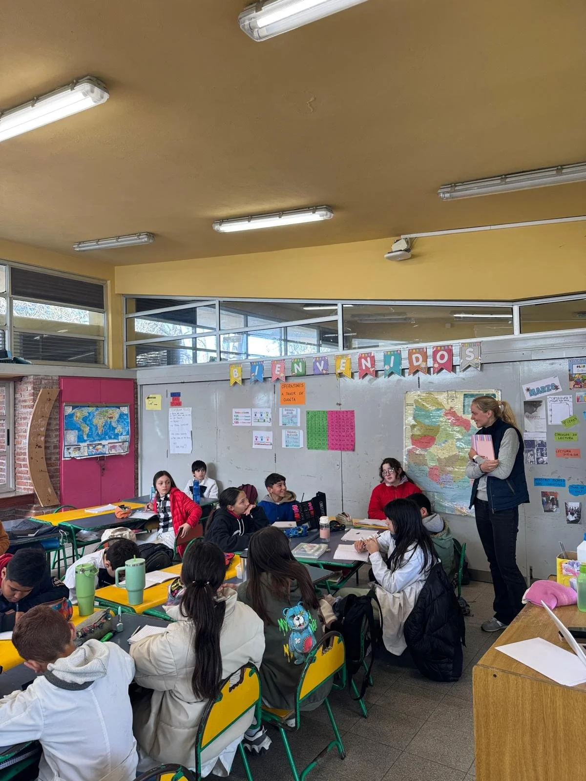 A classroom with students seated at colorful yellow and green desks, listening to a teacher standing at the front. The classroom has posters and maps on the walls, and a banner reading 'BIENVENIDOS' hangs above.