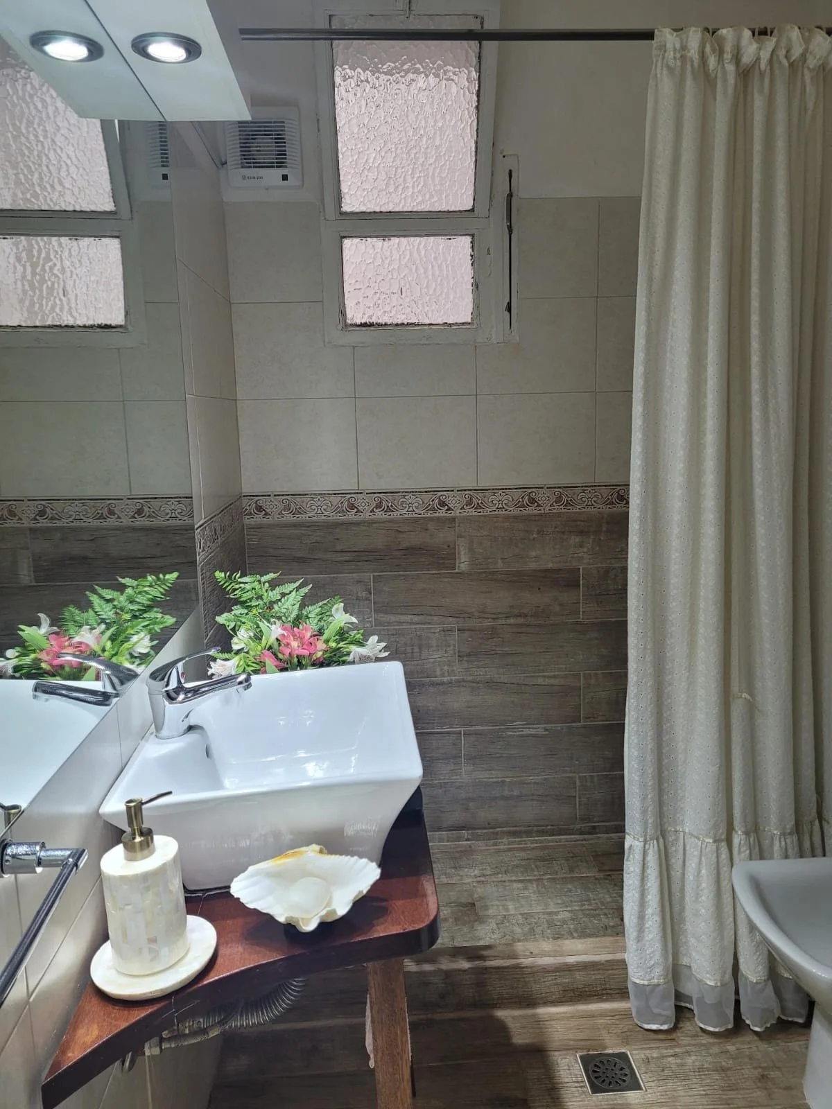 Bathroom with a white sink on a wooden vanity, a mirror, a soap dispenser, a shell-shaped dish, a fake plant, beige and brown tiled walls, frosted windows, a shower curtain, and a drain on the wooden floor.