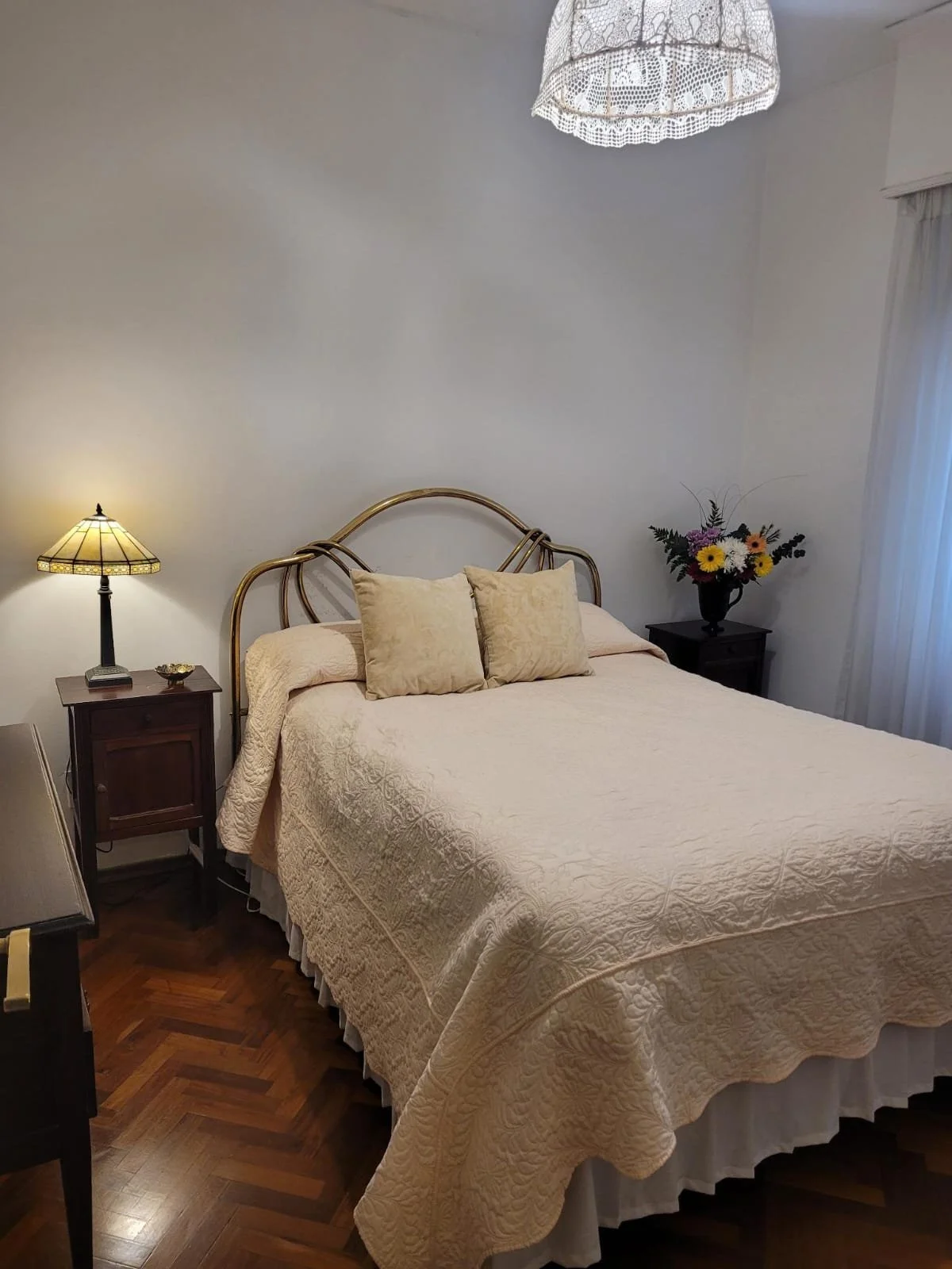 A cozy bedroom with a large bed covered in a cream quilt, two matching pillows, a wooden nightstand on each side, one with a vintage table lamp, the other with a vase of colorful flowers, and a lace ceiling light fixture.